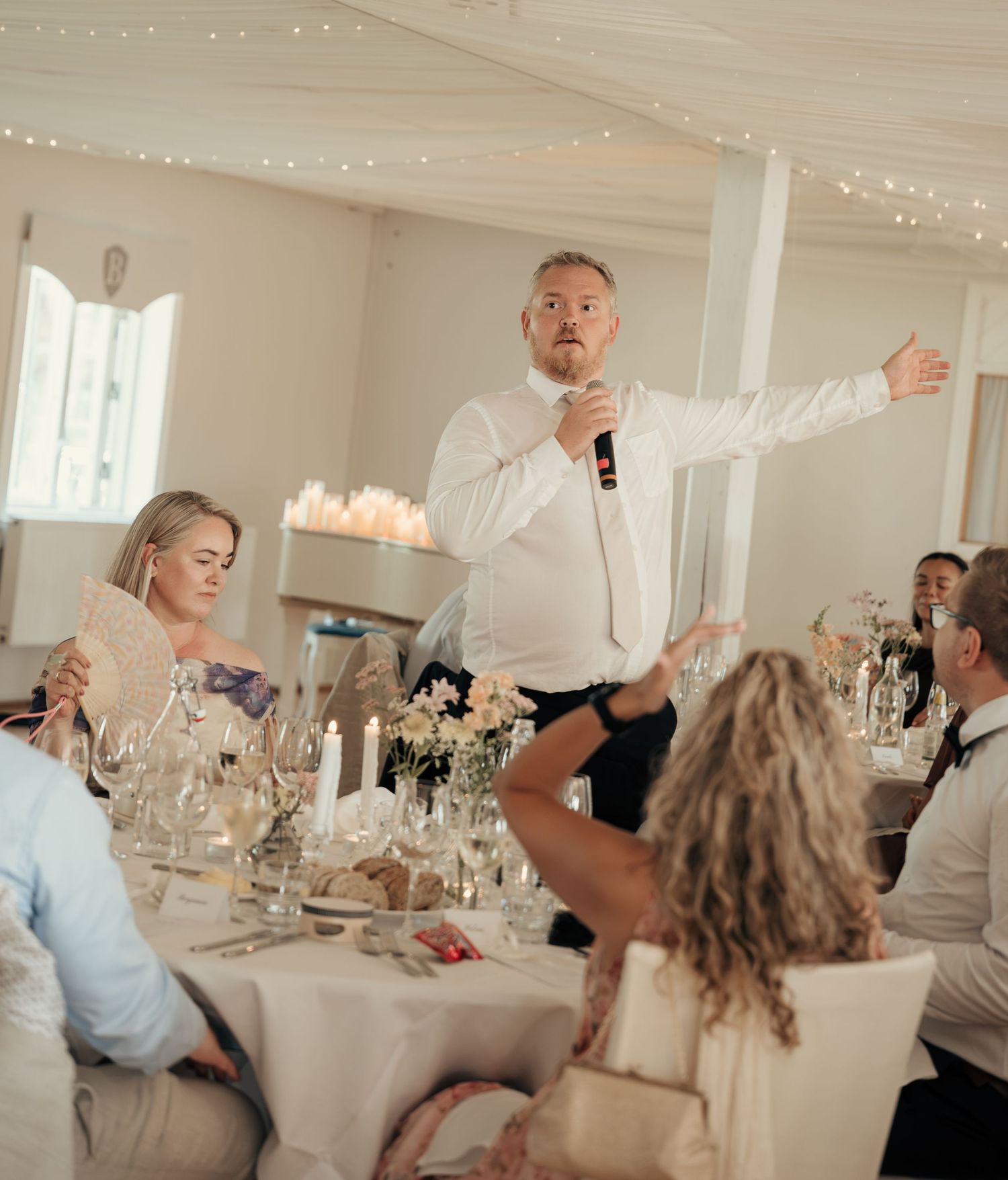 Man in a white shirt speaks into a microphone with an outstretched arm while seated wedding guests listen at a decorated reception table.