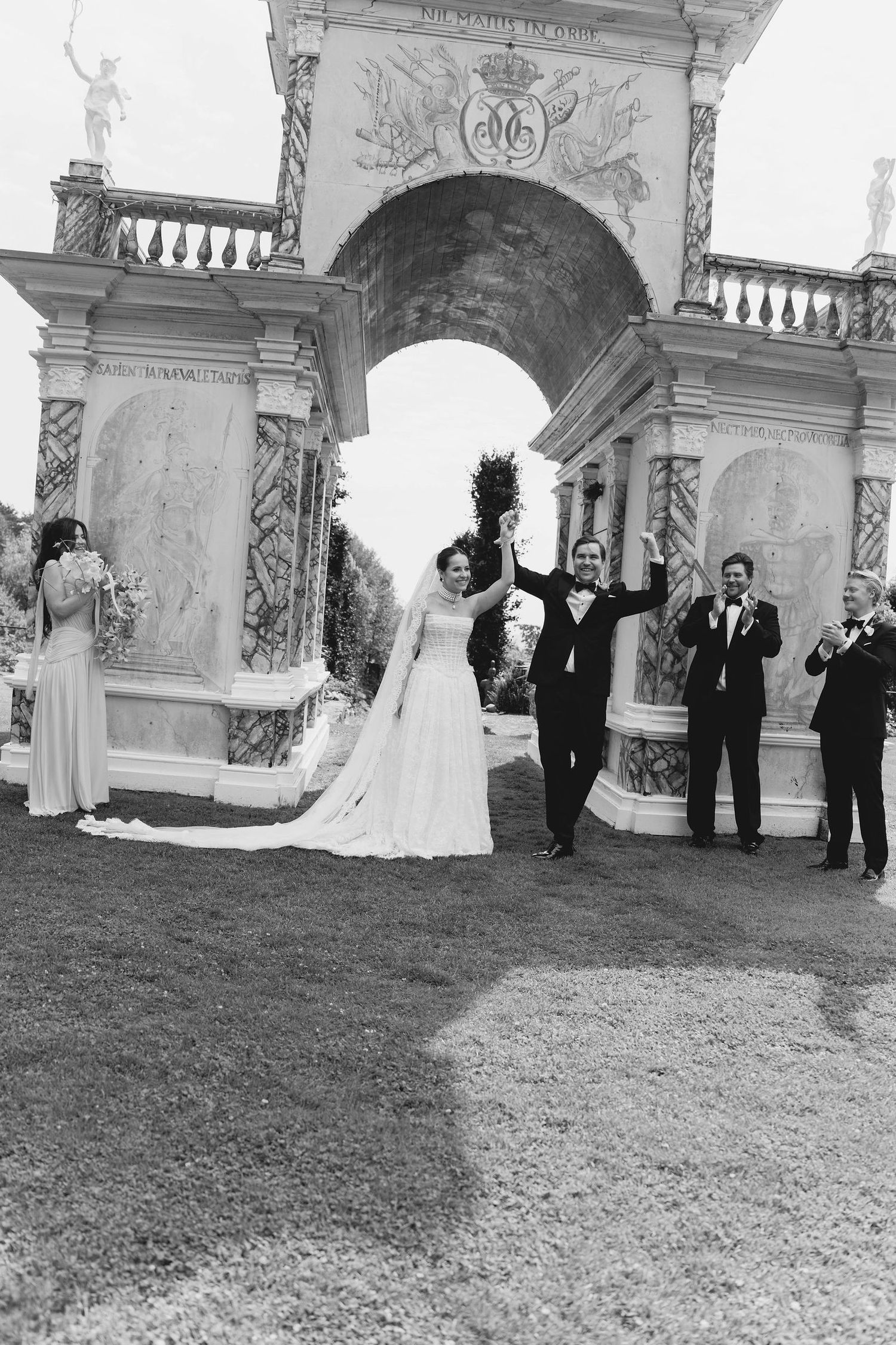 Black and white photo of a joyful bride and groom celebrating under an ornate arch at an elegant outdoor wedding ceremony.