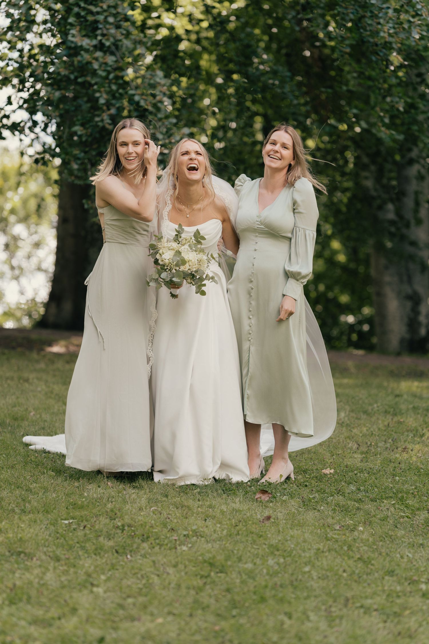 Bride laughing joyfully with two bridesmaids in sage green dresses outdoors on green grass.