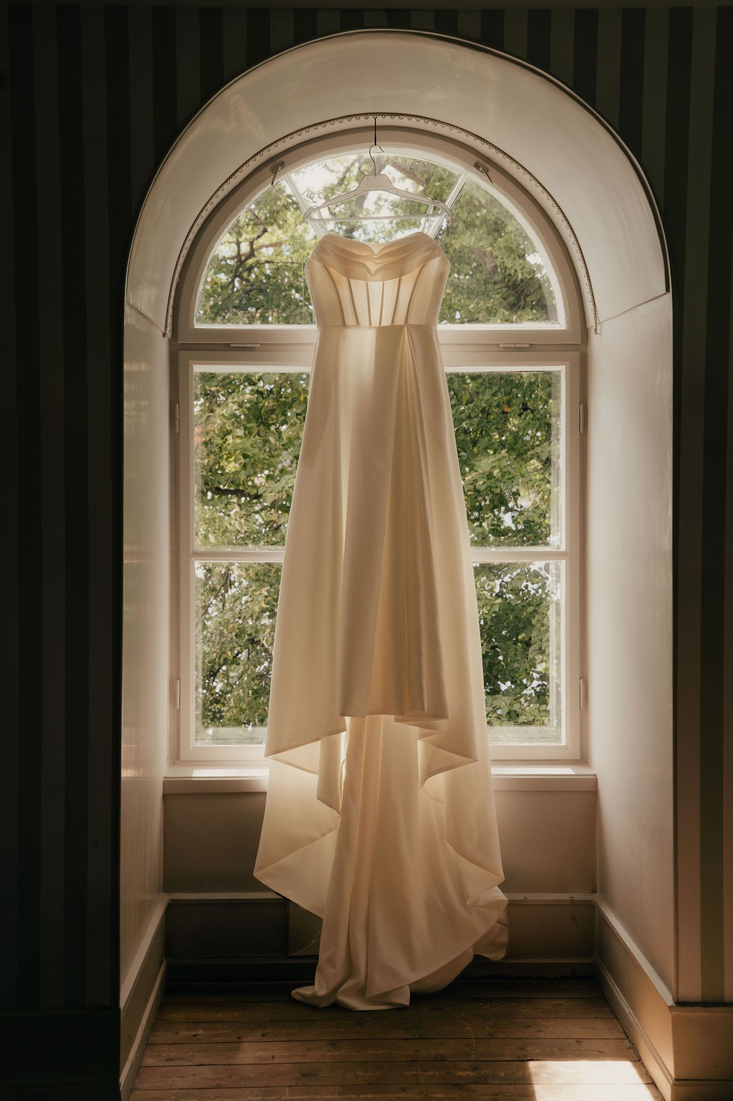 Elegant ivory wedding dress hanging in an arched window with natural light and lush green trees in the background.