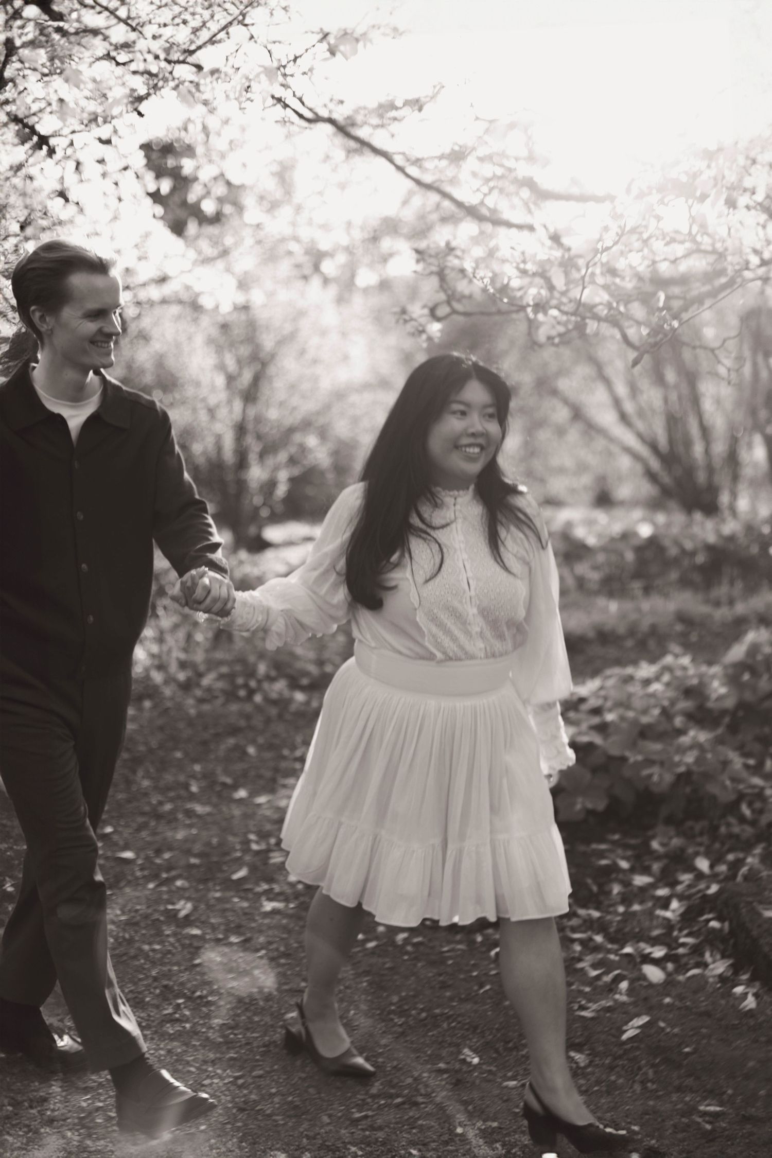 A couple walks hand in hand on a path under blooming cherry blossom trees in black and white photography.