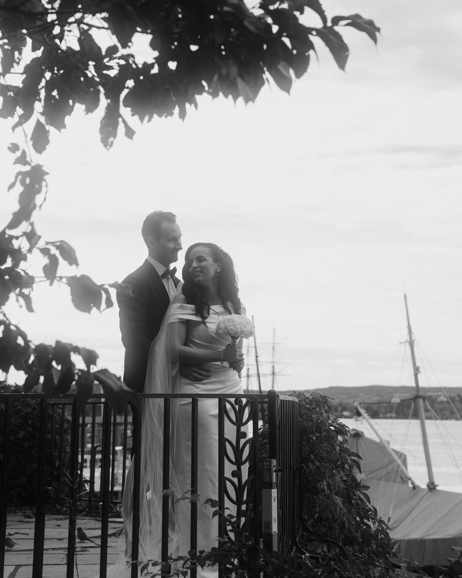 A couple embraces on a balcony overlooking water, framed by tree leaves in a romantic black and white photograph.