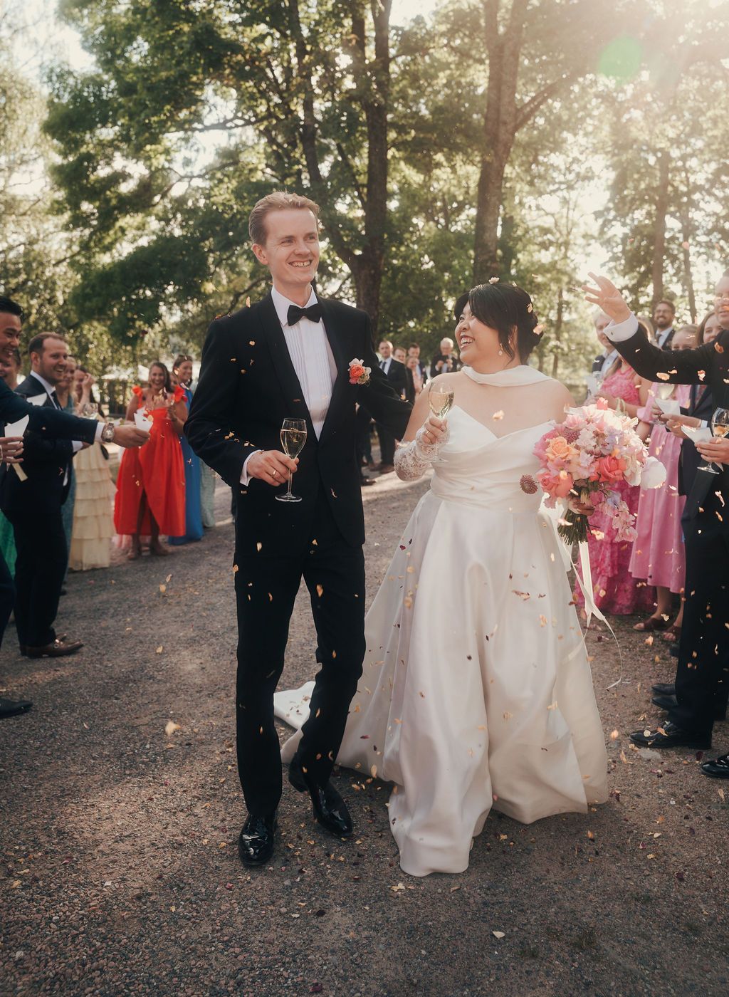 Wedding guests toss confetti and celebrate as newlyweds walk through an outdoor wooded area during sunset.