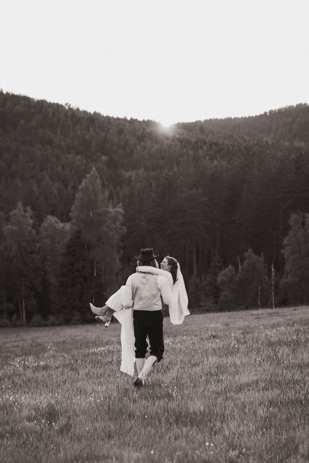 A couple in formal attire walks together through a scenic mountain meadow in black and white photography.