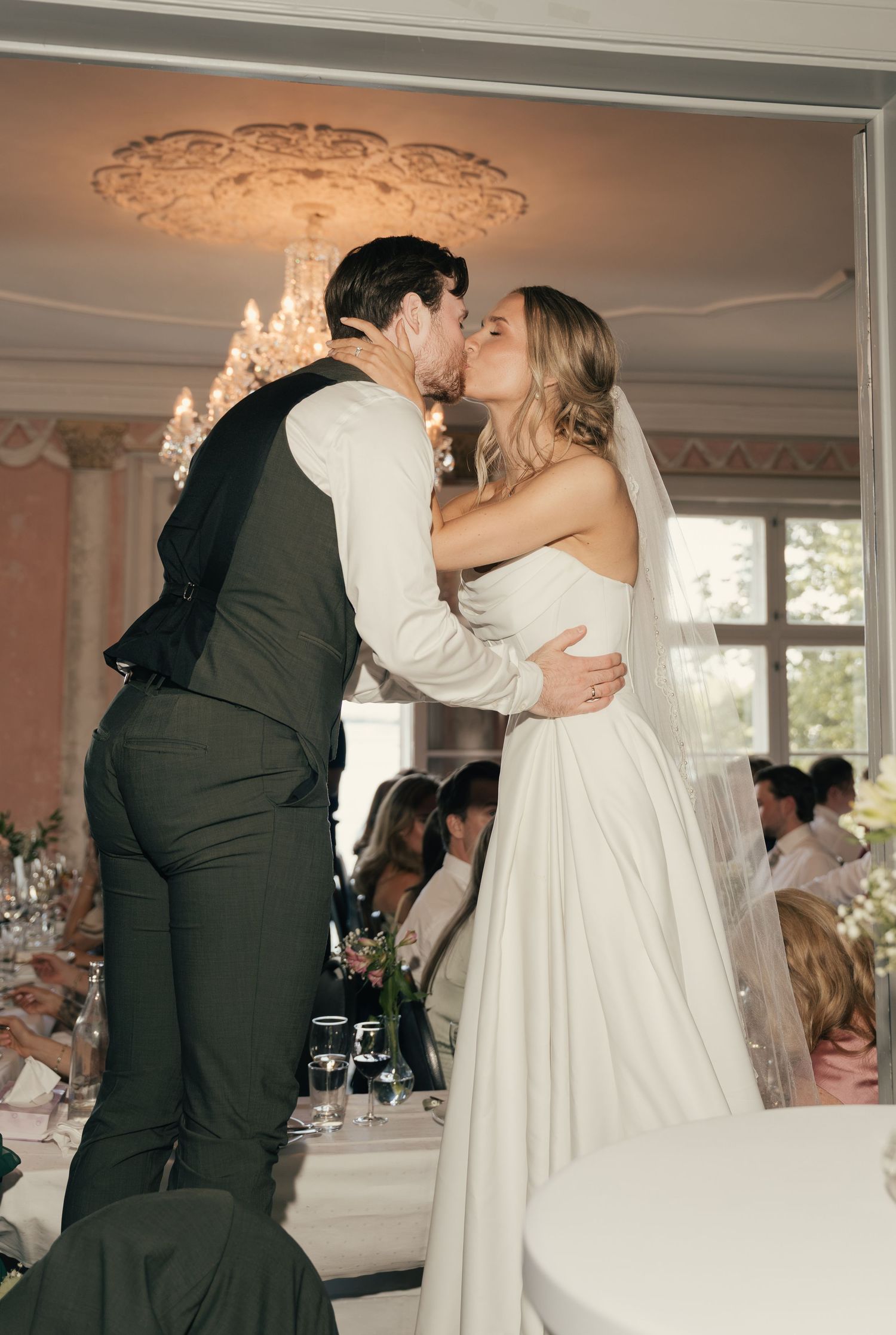 Newlyweds sharing first kiss at elegant indoor wedding reception with chandelier and guests seated.