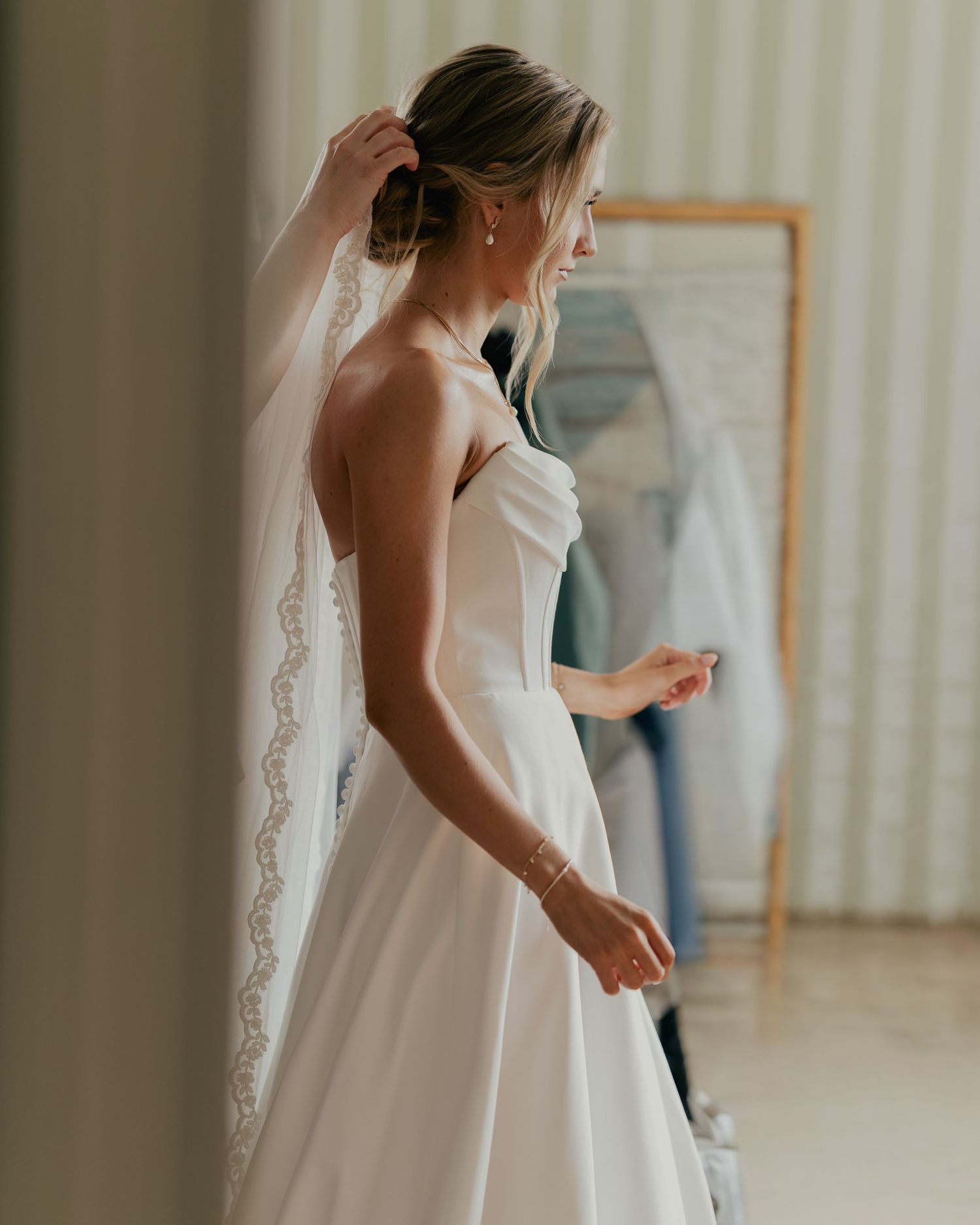 Bride in strapless white wedding gown and lace veil, elegant updo hairstyle, gold bracelet, getting ready indoors.