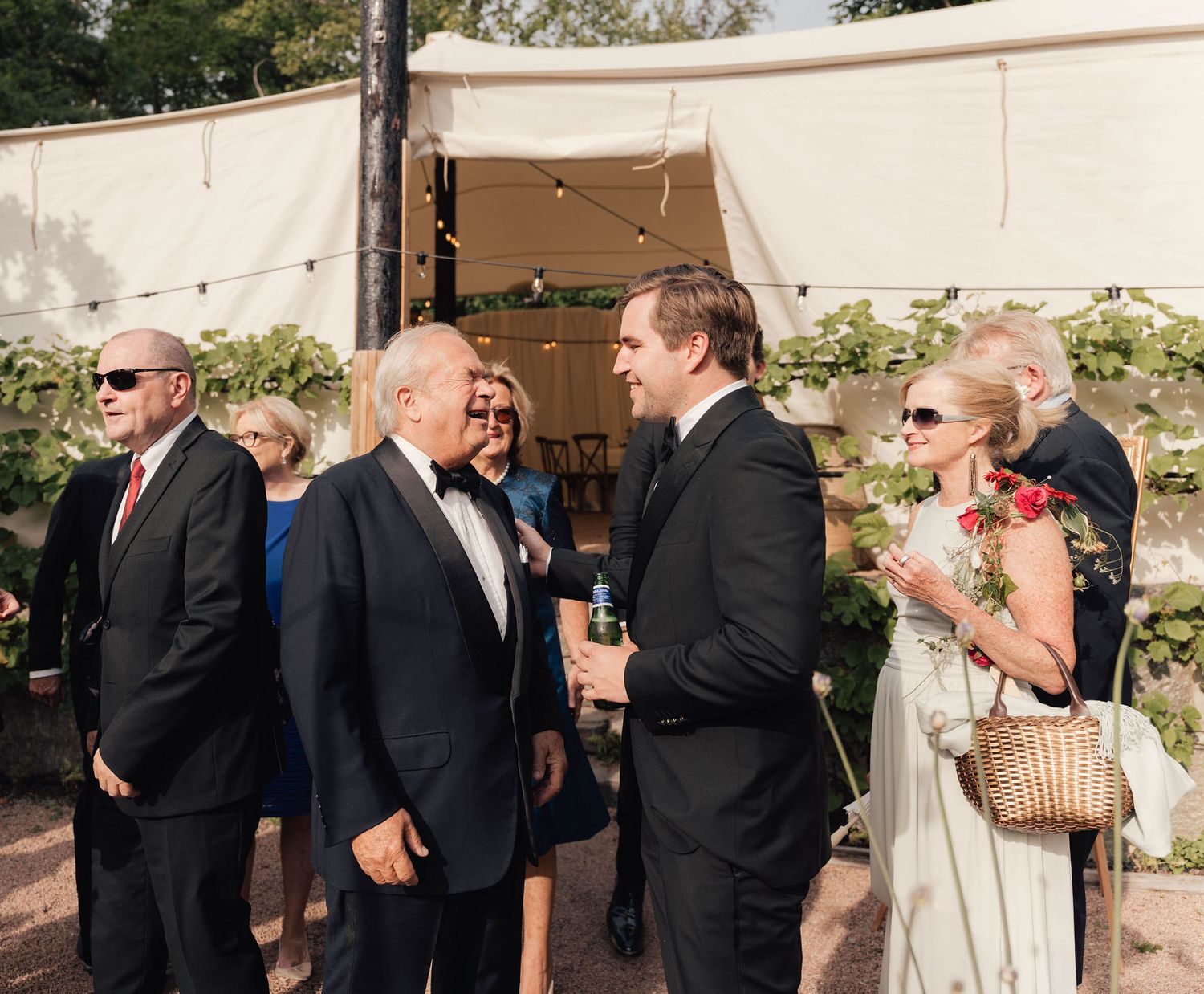 Elegantly dressed wedding guests socializing outdoors near a white tent decorated with string lights and greenery.