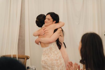 Two women share a heartfelt hug at a wedding reception, one wearing a lace bridal gown, with guests applauding in the foreground.