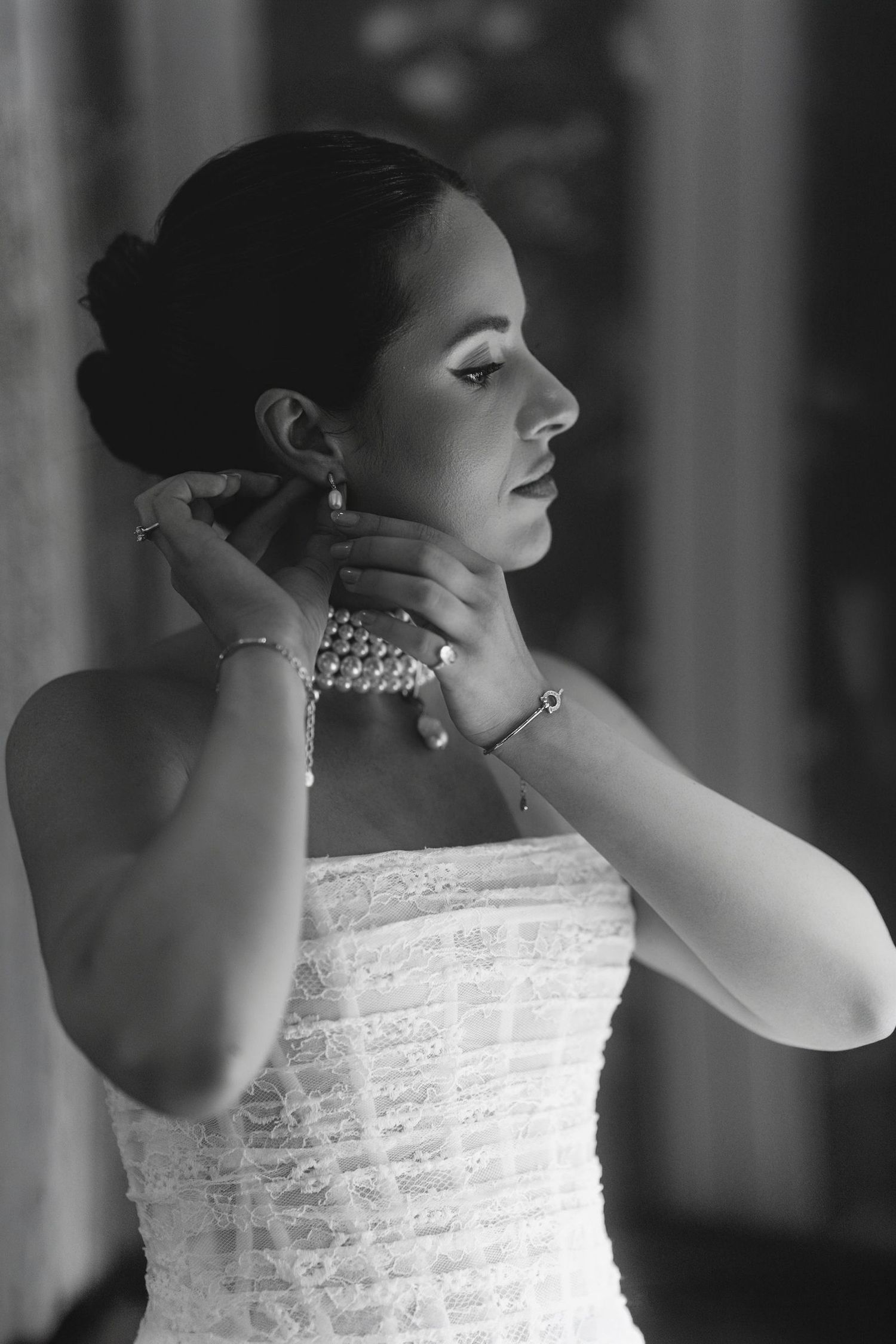 Black and white photo of a bride in a strapless lace gown adjusting her pearl earring and bracelet.