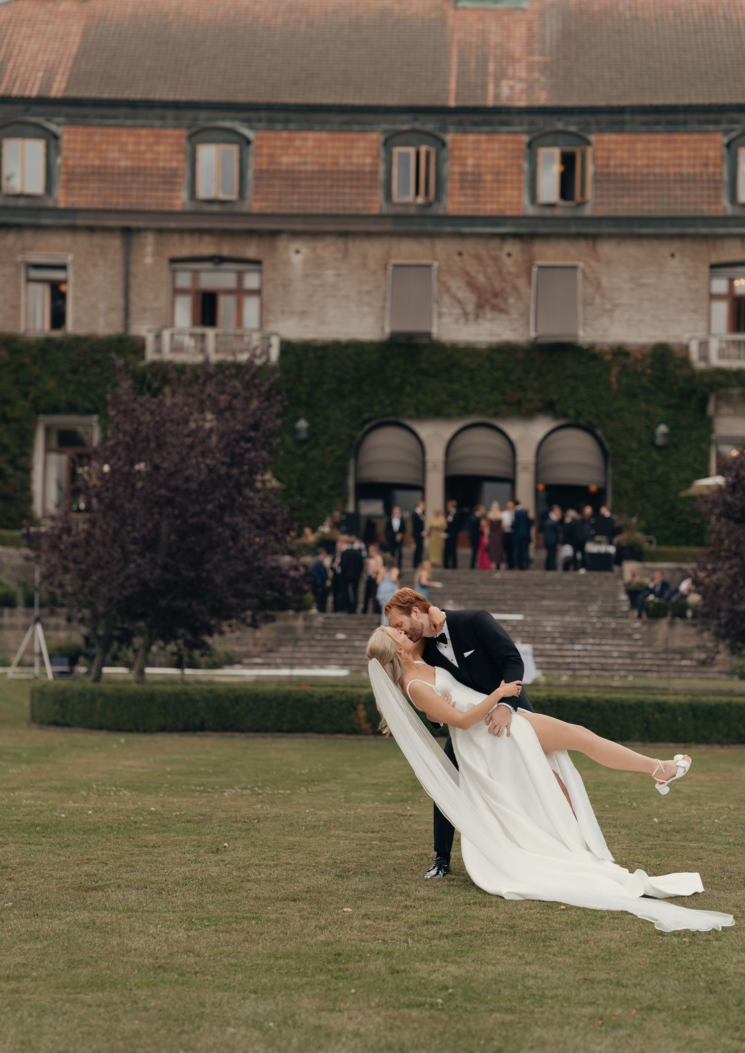 Groom dips bride in a white gown on a manicured lawn before a historic ivy-covered estate as wedding guests gather on the steps.