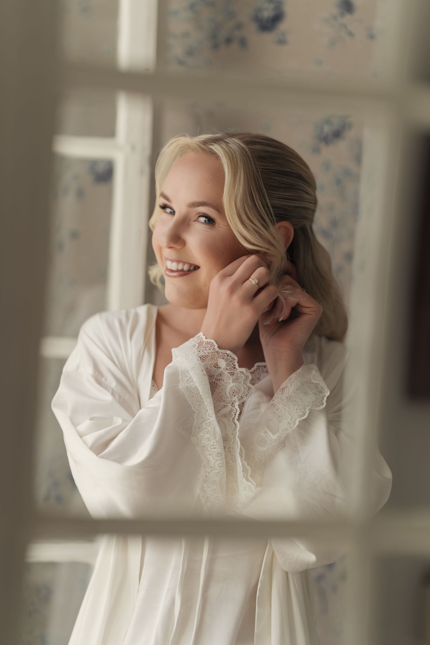 Smiling bride with blonde hair in a white lace-trimmed satin robe putting on her earring while getting ready on her wedding day.