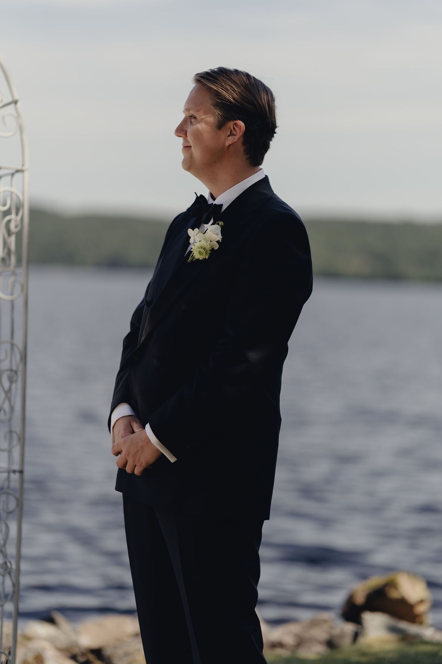 Groom in a black tuxedo with a white floral boutonniere stands waiting beside a lakeside wedding arch.