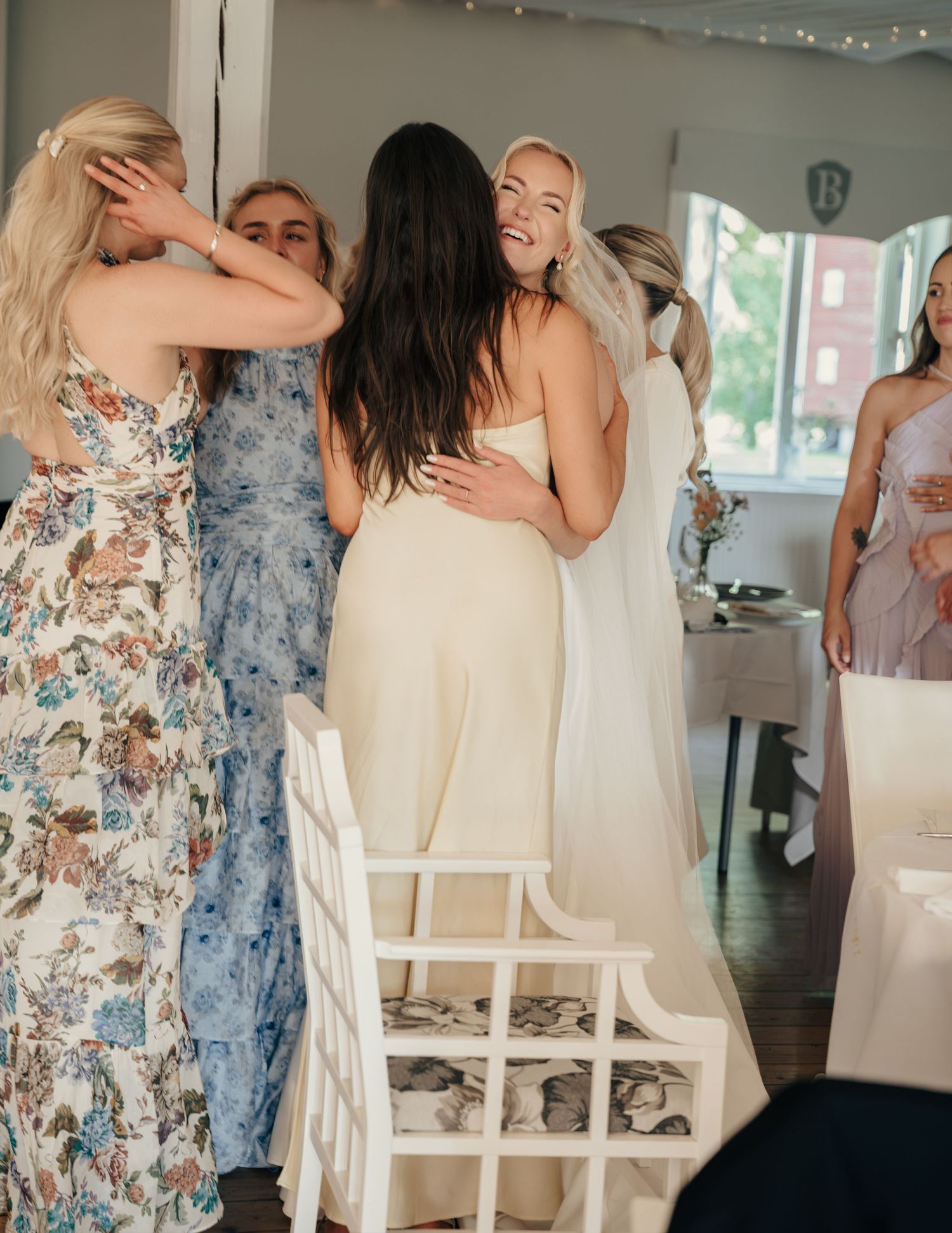 Smiling woman in a cream satin dress hugs a bride in white as bridesmaids in floral gowns gather in a bright reception room.