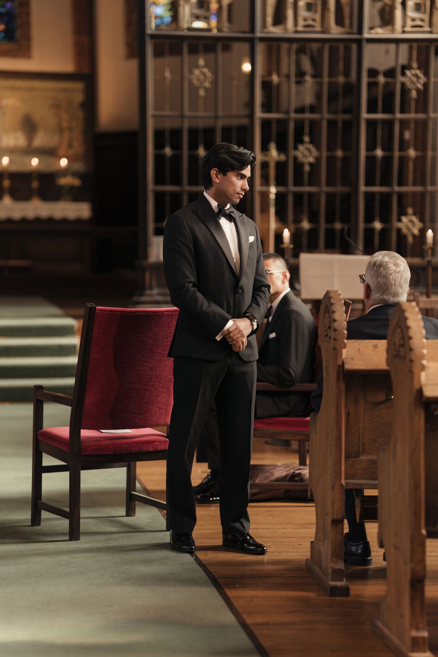 Groom in a black tuxedo and bow tie stands nervously at the front of a church, with guests seated in wooden pews behind him.