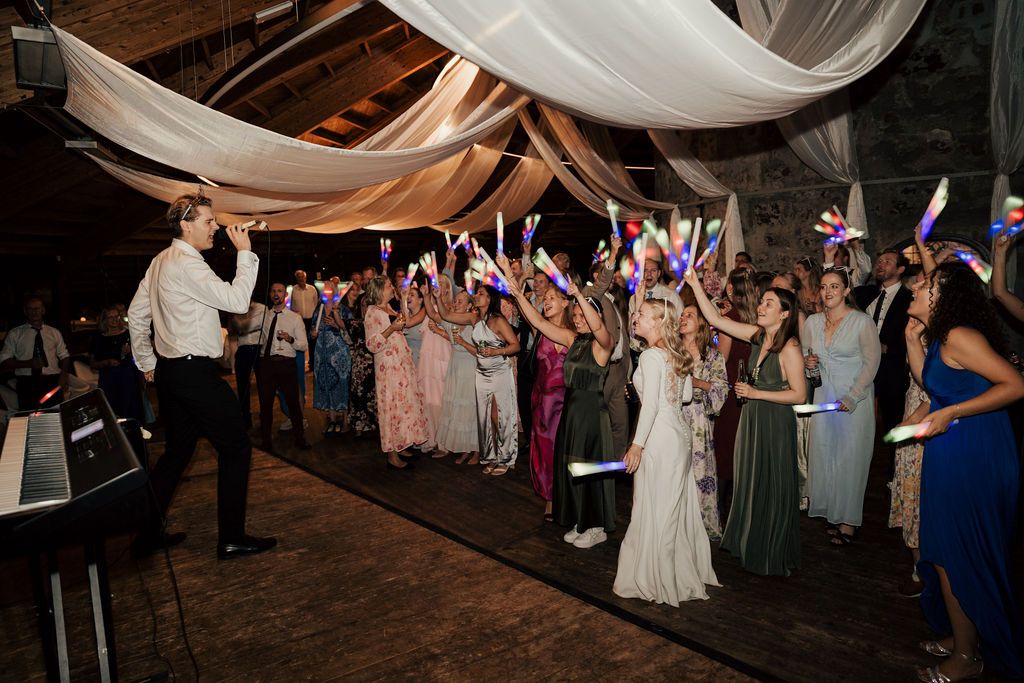 Wedding reception crowd dancing and celebrating with arms raised under draped fabric decorations at a rustic venue.