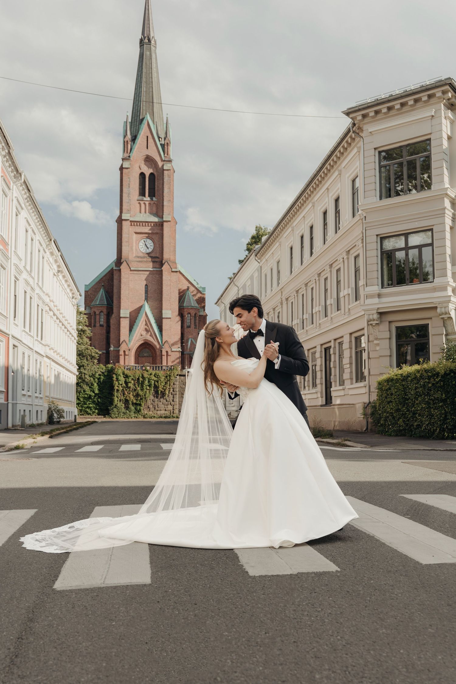 Bride in a white gown with a long veil and groom in a tuxedo share a dance on a crosswalk, with a red-brick church tower behind them.