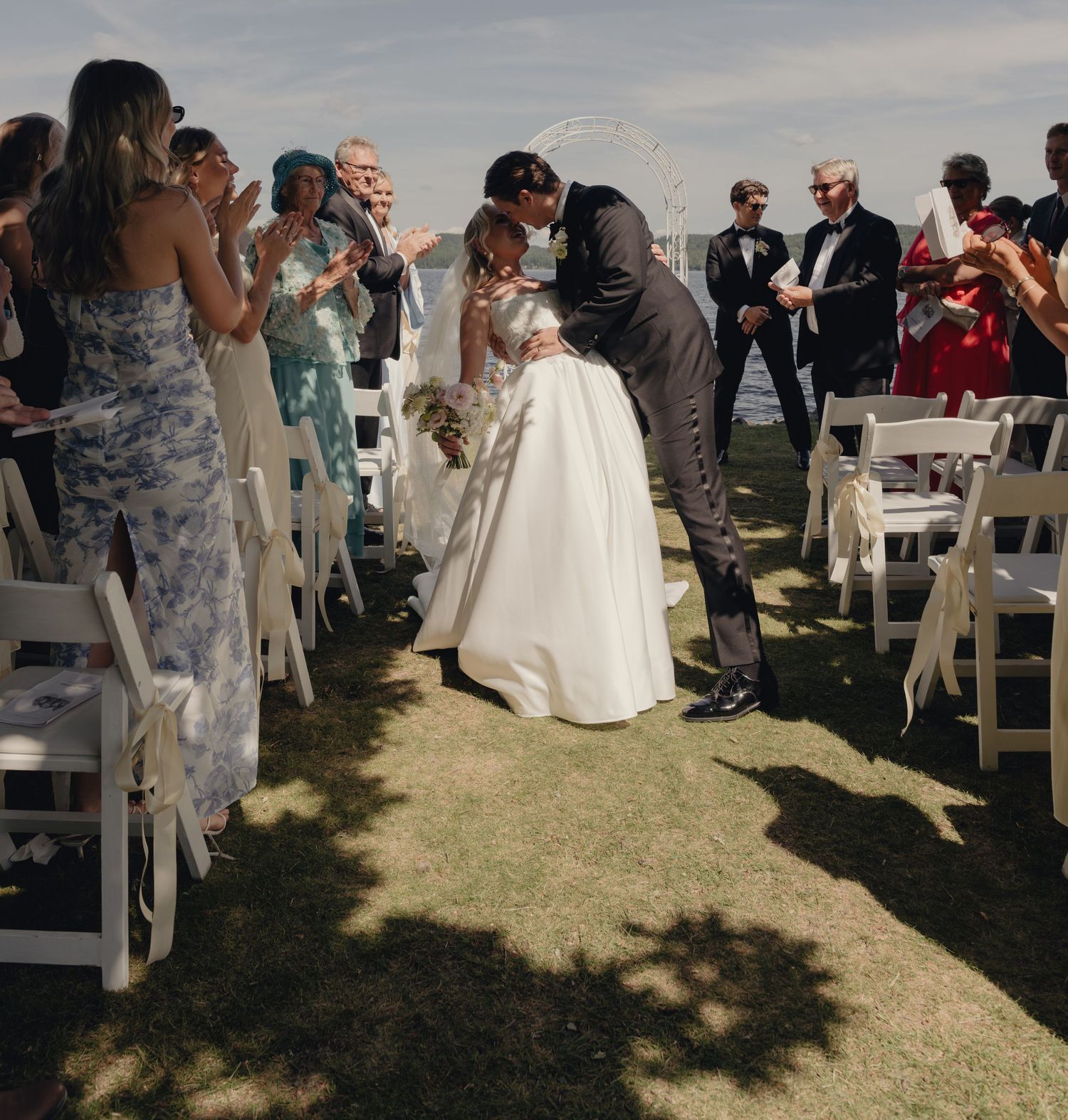 Bride in a white gown and groom in a dark suit kiss on a grassy aisle as wedding guests cheer from white chairs at an outdoor ceremony.