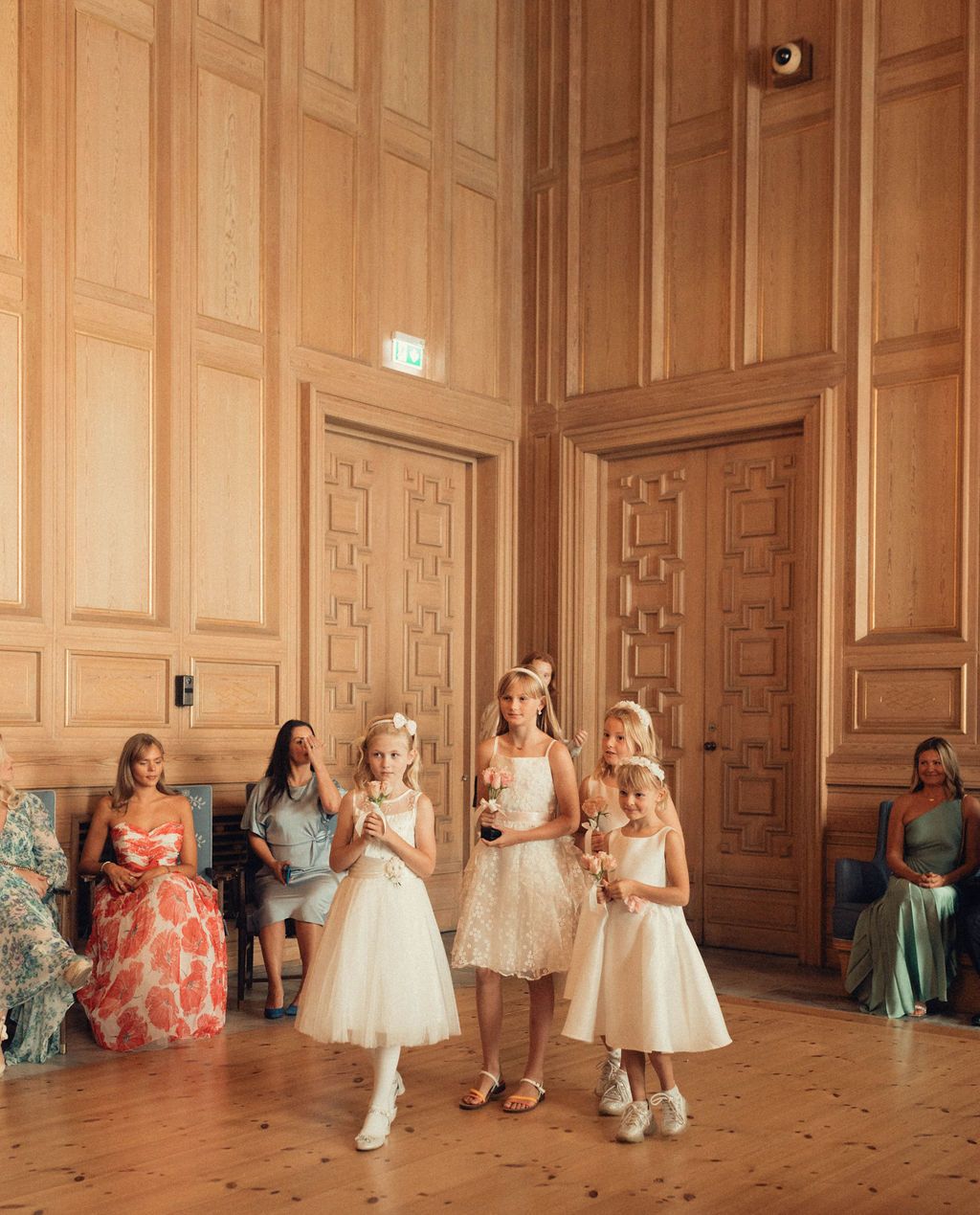 Young girls in elegant party dresses dance and twirl in a grand ballroom with ornate wood-paneled walls.