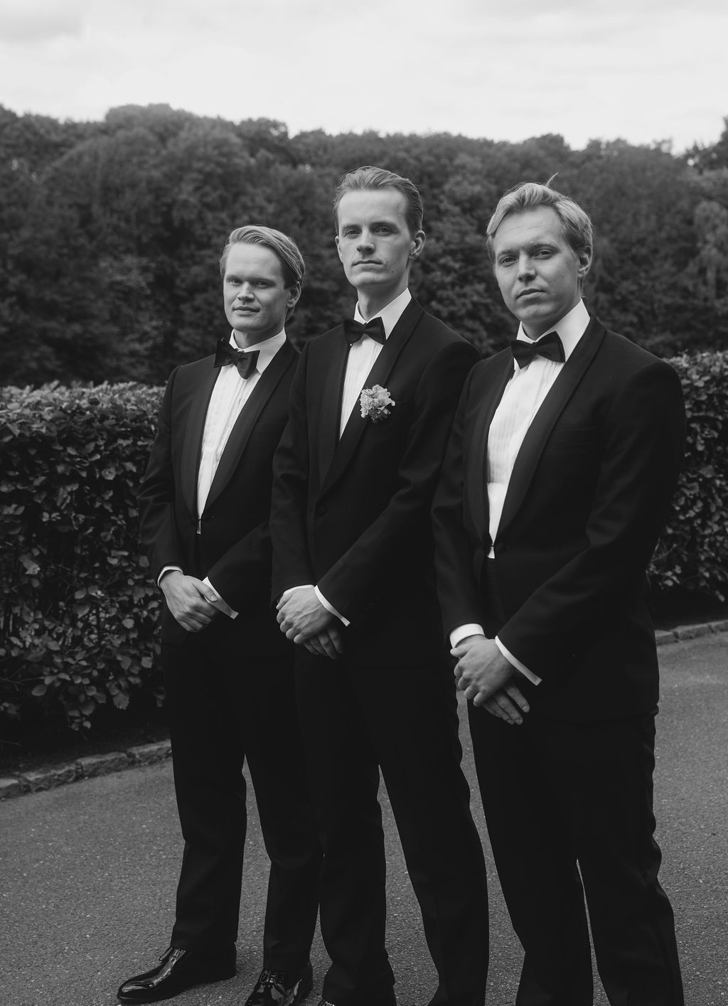A group of men in formal black tuxedos and bow ties stand together outdoors in a black and white photograph.