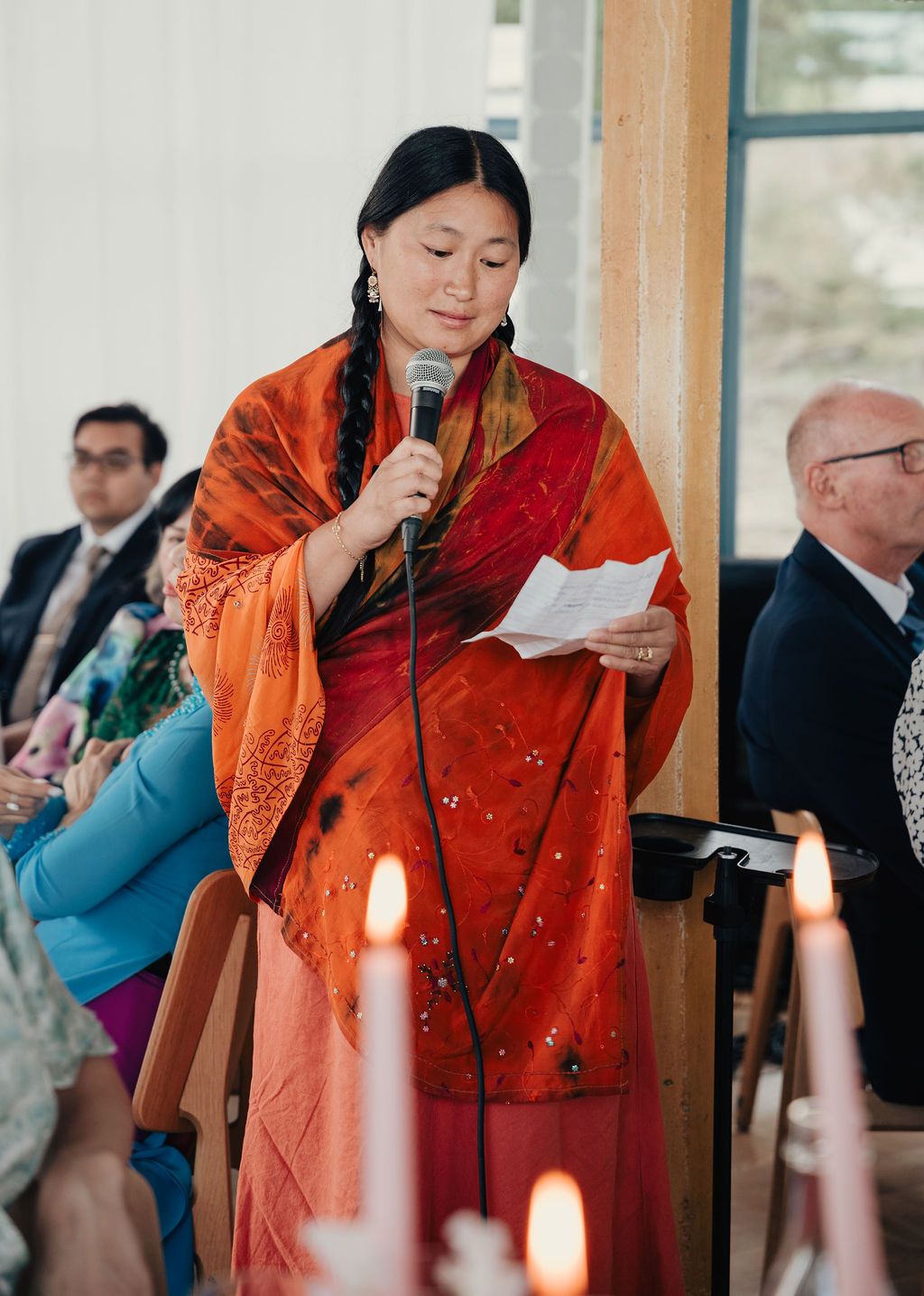 A person wearing vibrant orange and red traditional attire reads from papers during an indoor event.
