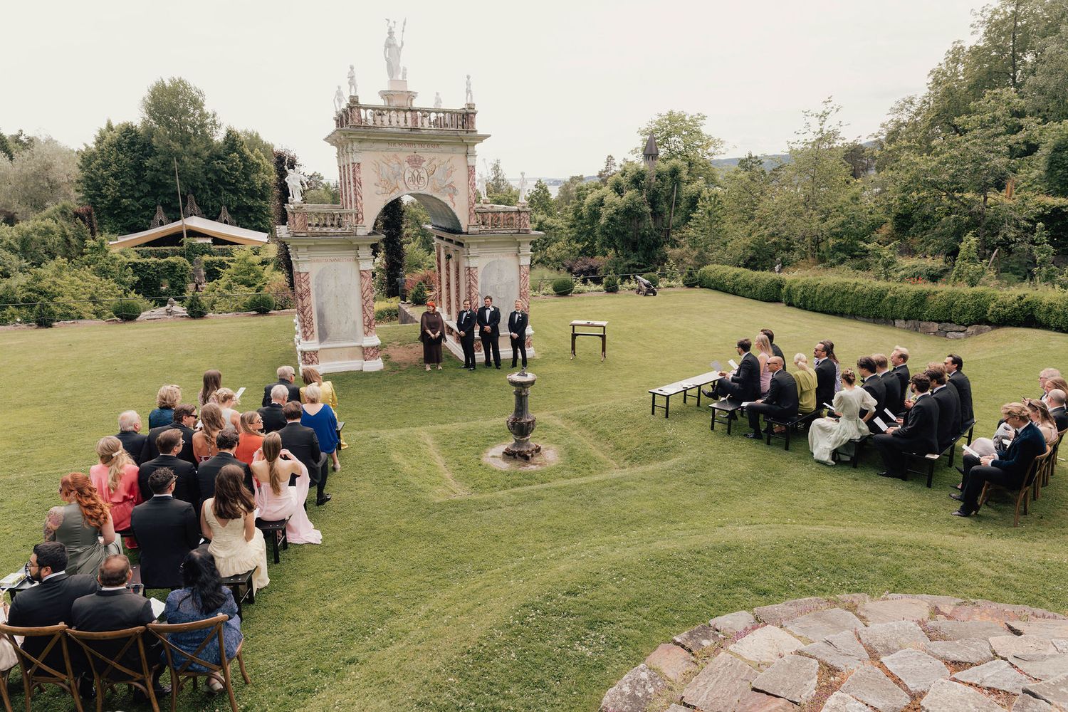 Outdoor wedding ceremony in a lush garden with guests seated on benches facing an ornate stone archway.