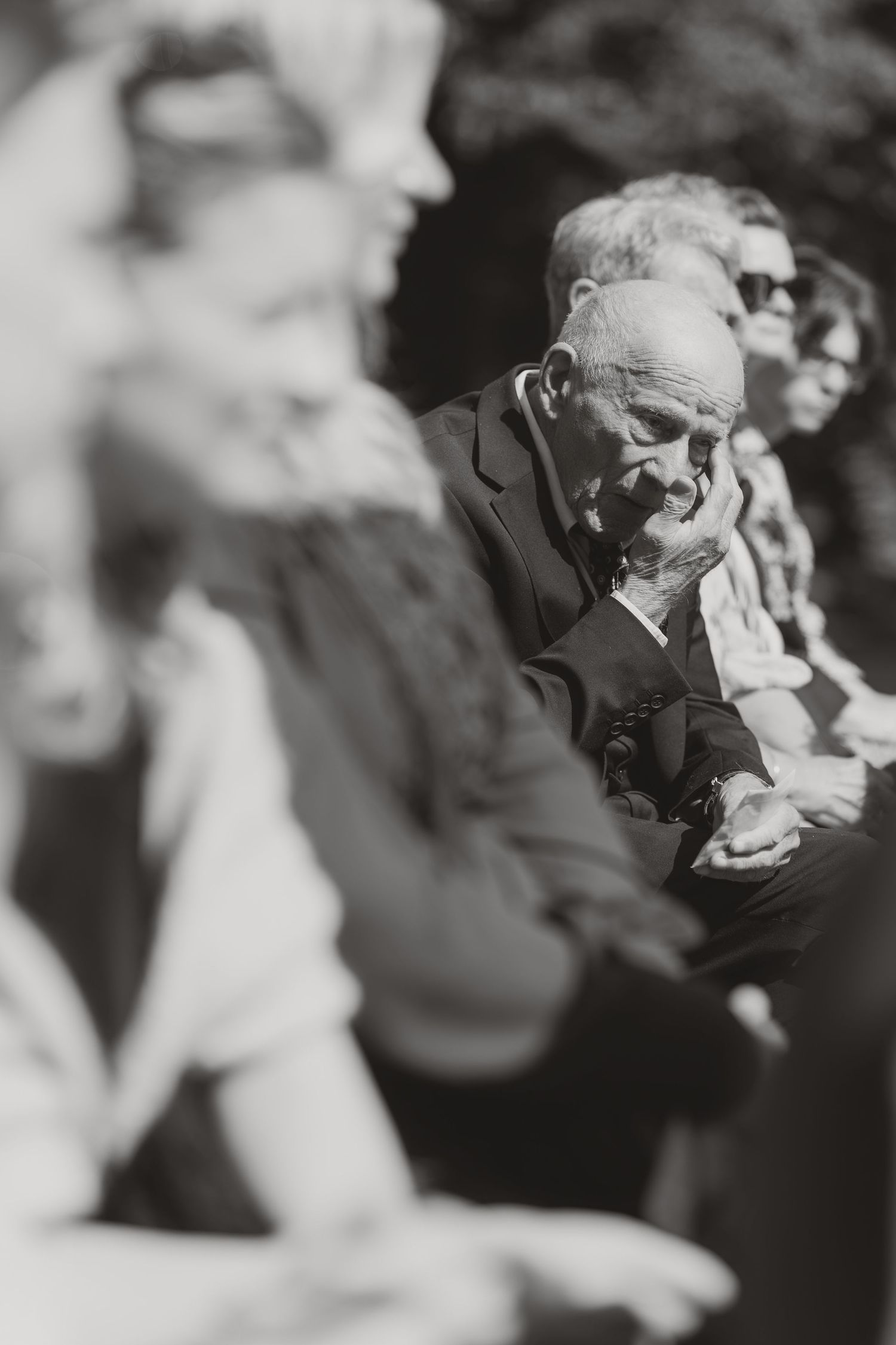 Black and white photo of elderly man resting chin on hand, seated among blurred figures in an outdoor gathering.