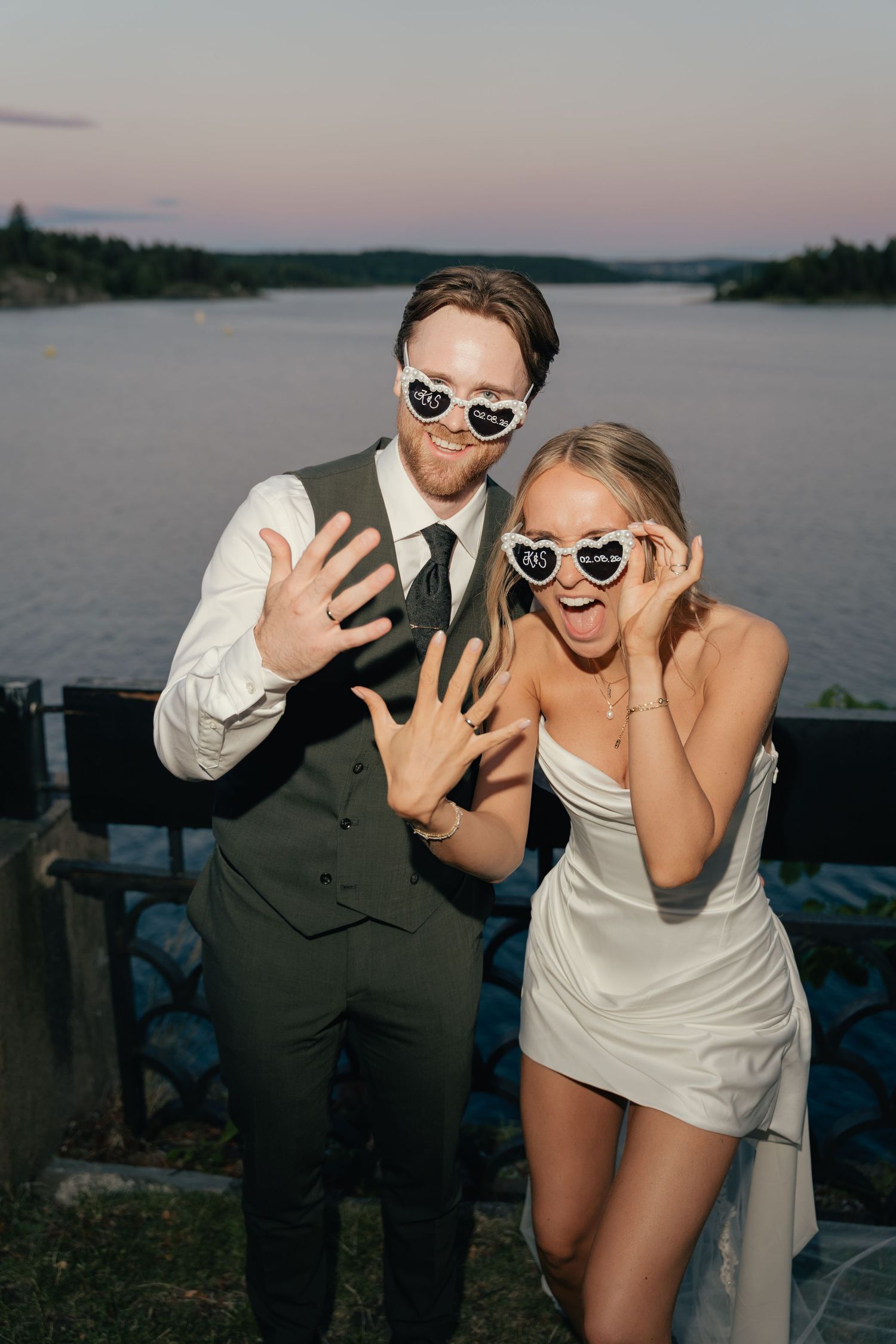 Newlyweds wearing heart-shaped sunglasses posing playfully by a scenic lakeside at sunset.
