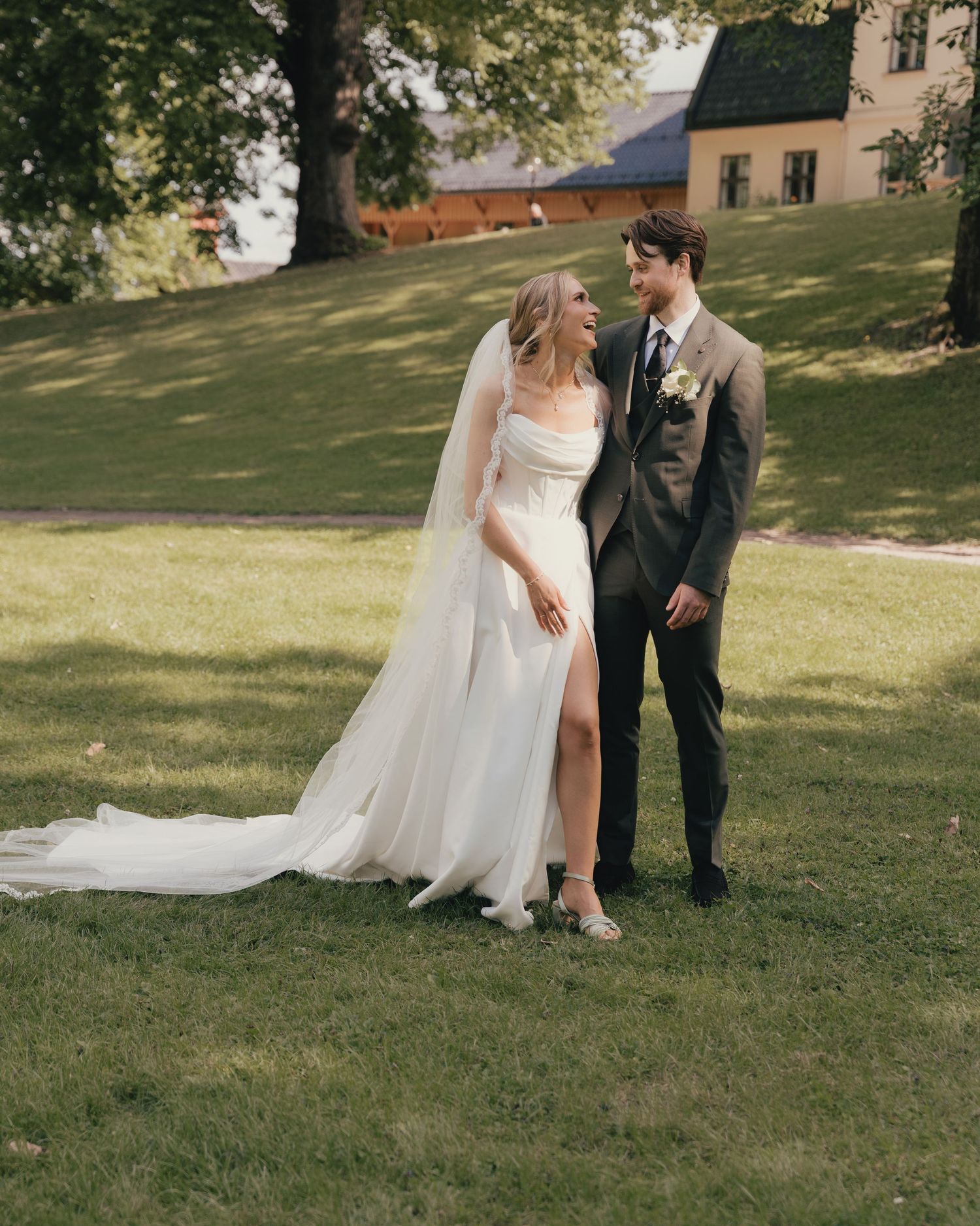 Bride and groom share a joyful moment on green lawn, bride in strapless white gown with veil and slit.