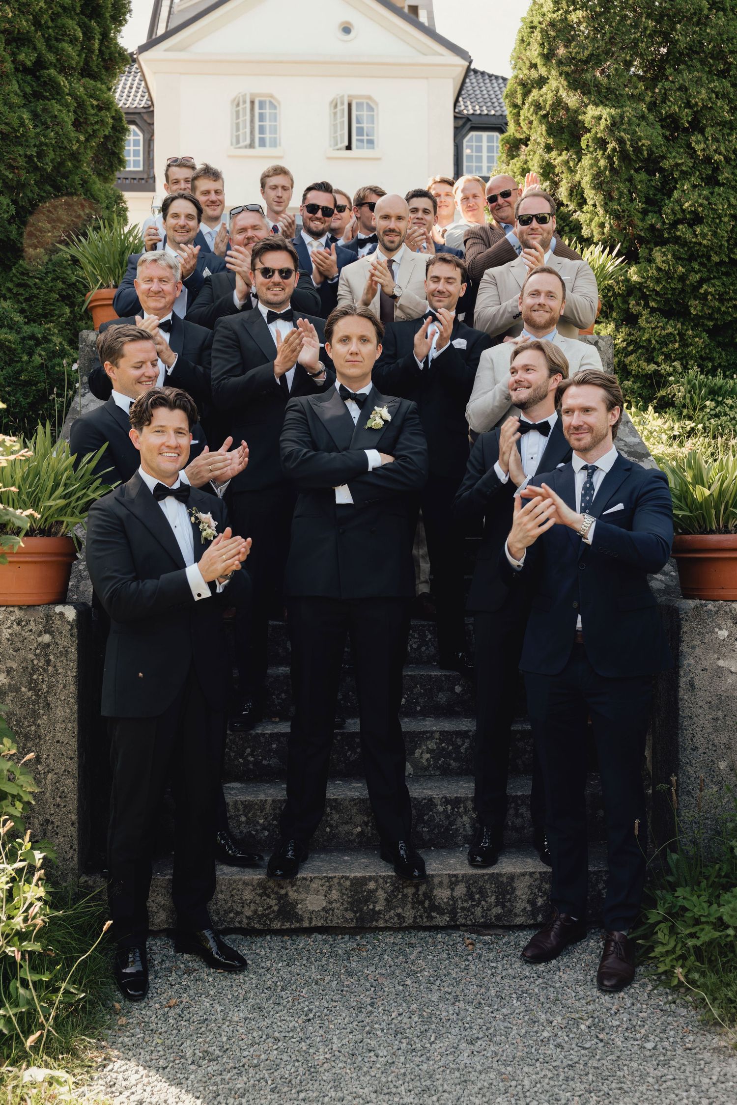 A group of groomsmen in dark suits and bow ties applaud on stone steps outside a historic white building on a sunny day.