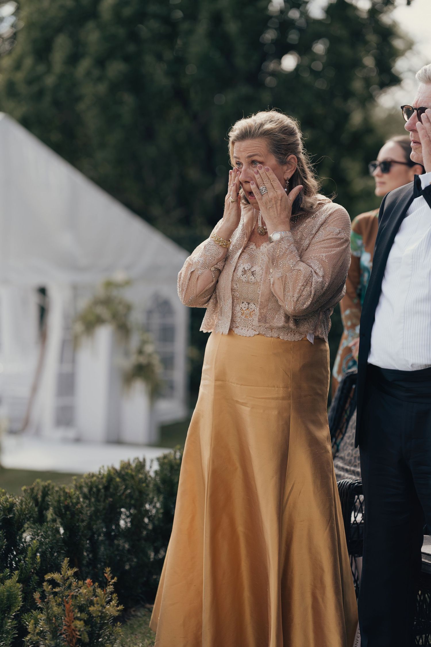 Emotional woman in a gold satin skirt and lace blouse covers her face with her hands at an outdoor wedding with a white tent behind.