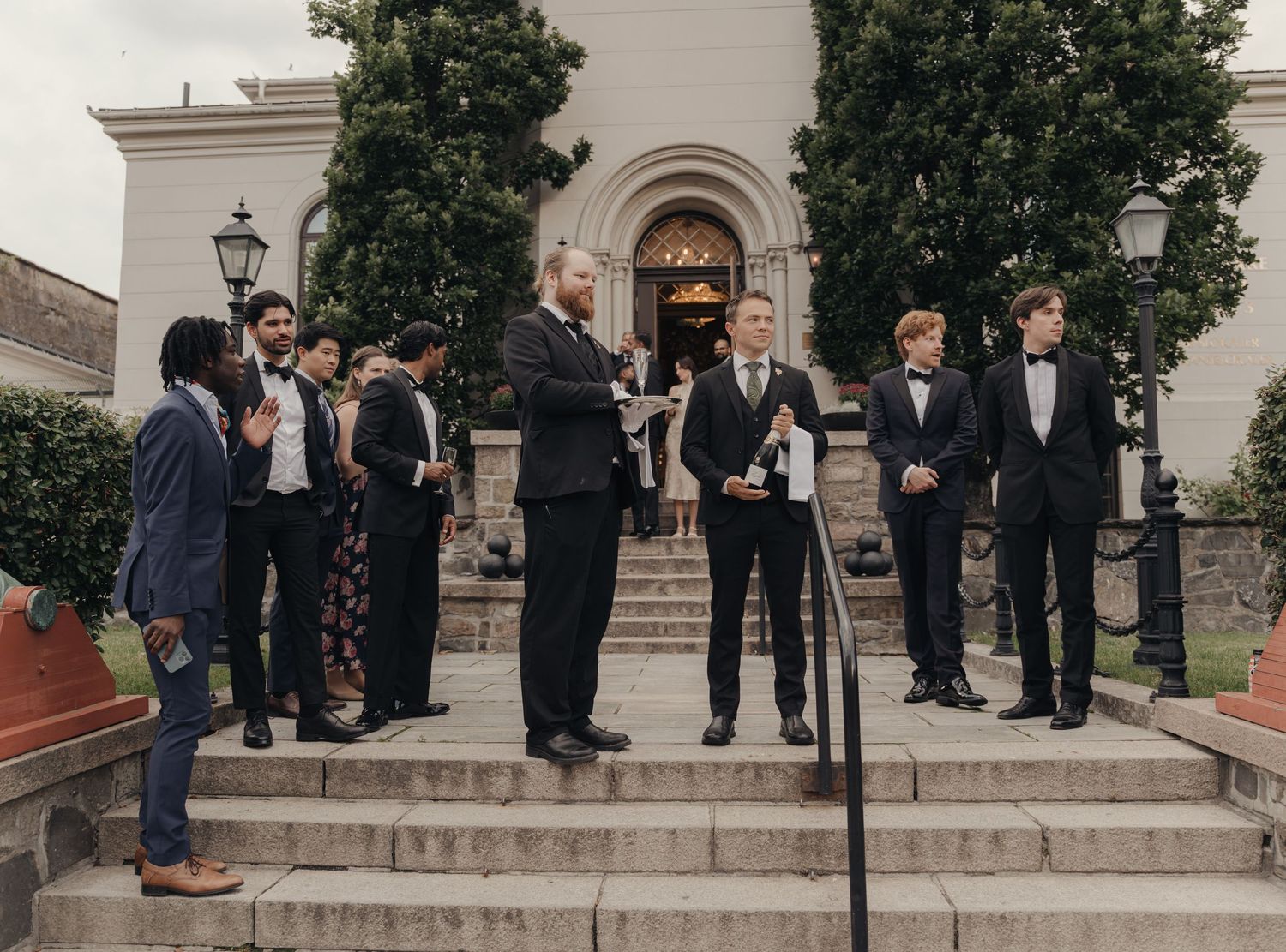Group of groomsmen in dark suits and bow ties standing on stone steps outside a historic building with an arched entrance on a wedding day.