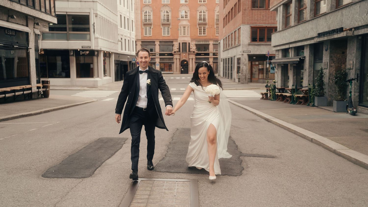 A couple in formal wedding attire runs joyfully down an empty city street together on their special day.