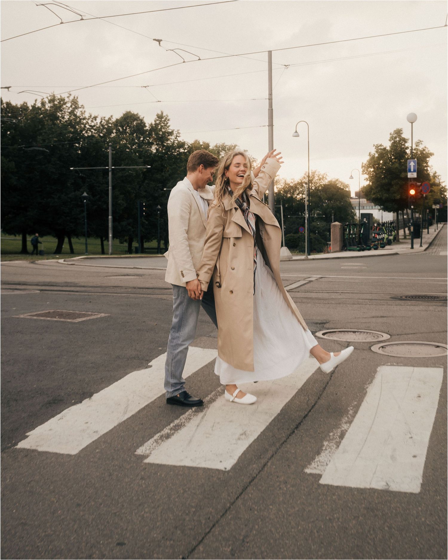 Two people in beige coats share a tender moment while crossing a street at a crosswalk during sunset.