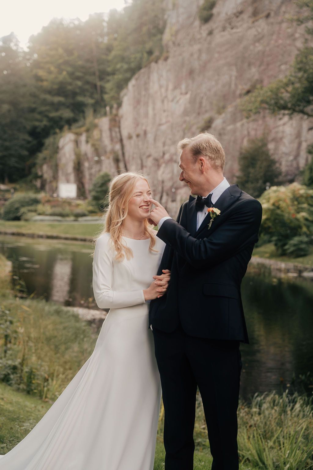 A couple shares joyful moments during their wedding photoshoot in front of a scenic rocky cliff and lake backdrop.
