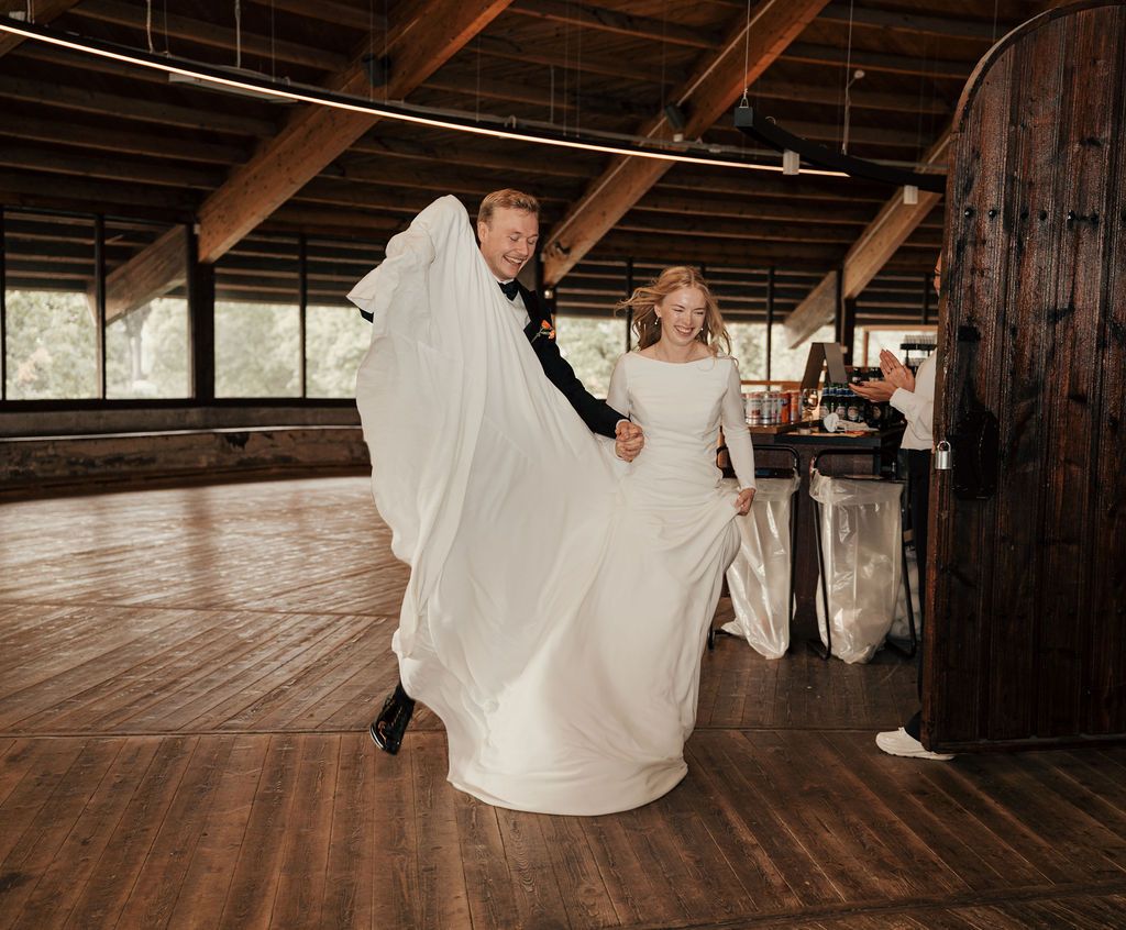 Joyful couple twirls and dances together on a rustic wooden floor in a barn-style venue with exposed beams.