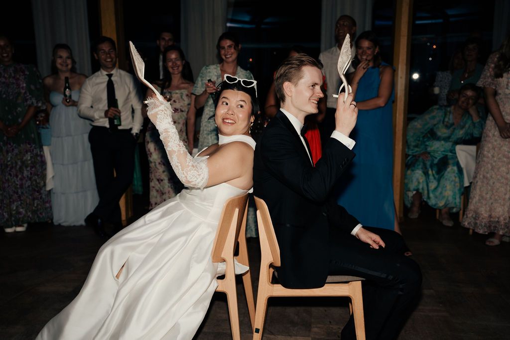 Wedding couple plays a fun game during reception while seated back to back in chairs with white gloves.