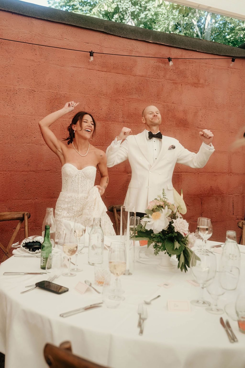 A joyful couple in wedding attire dance and celebrate at their reception table decorated with floral arrangements.