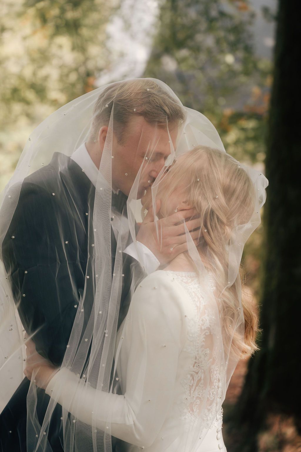 A romantic wedding couple shares an intimate moment under a veil in a sun-dappled outdoor setting.