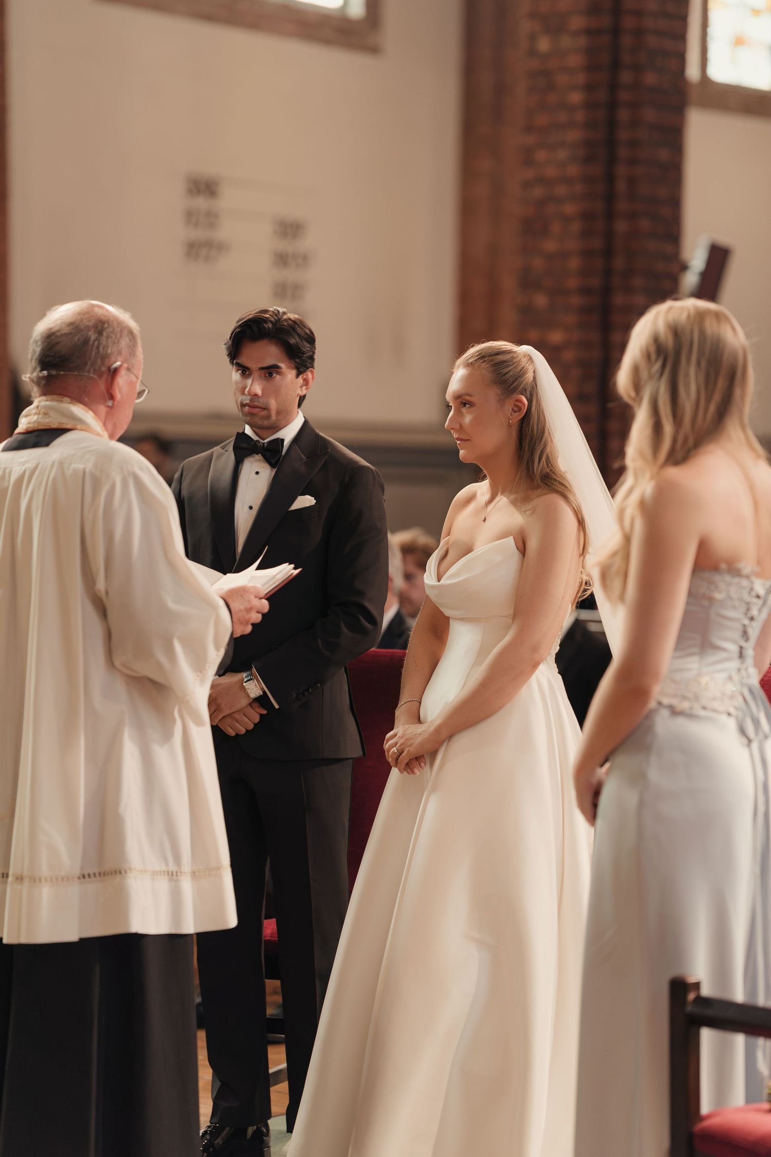 A bride in a strapless white gown and veil stands with her groom in a tuxedo as an officiant leads the church wedding ceremony.