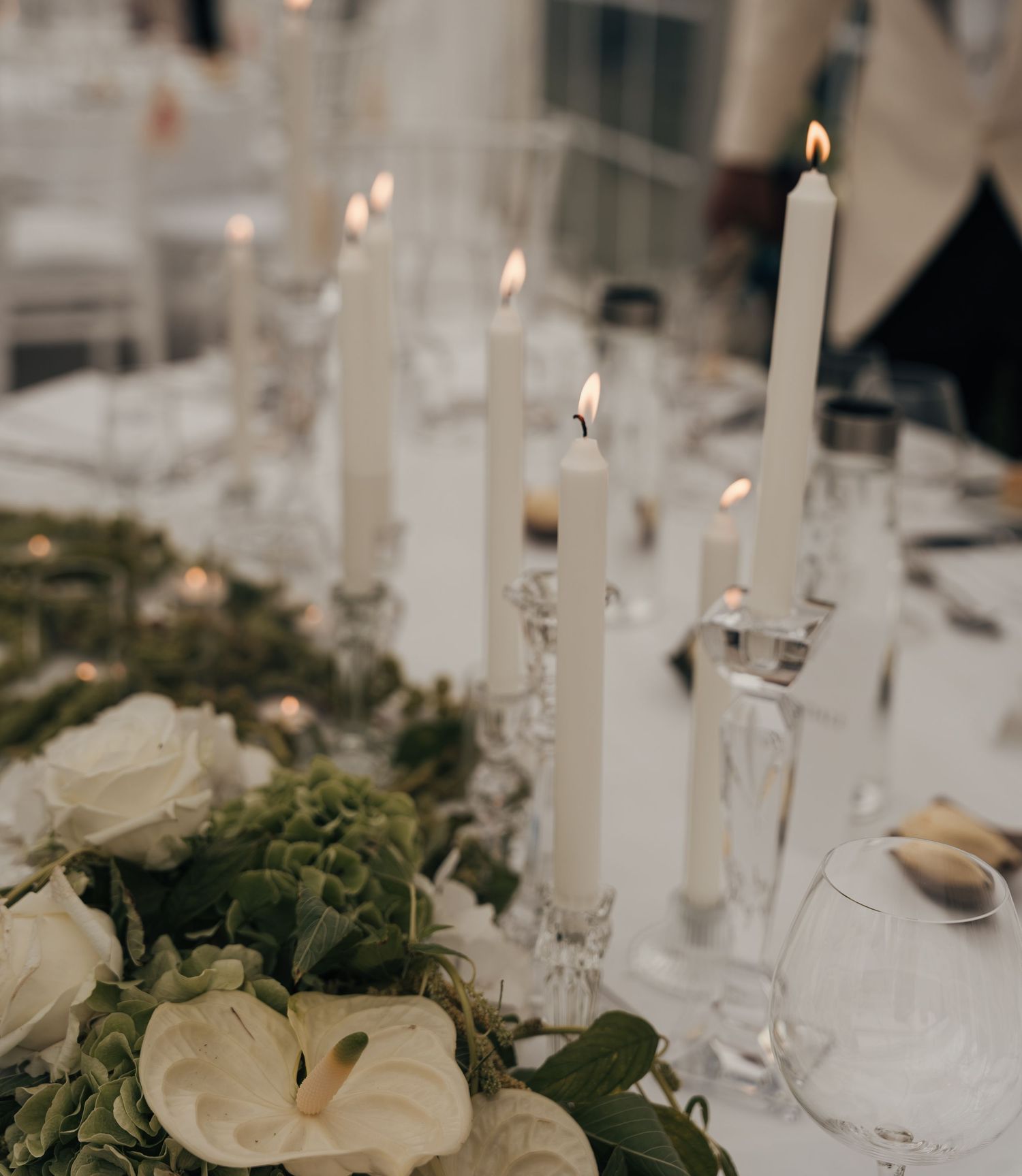 Lit white taper candles in glass holders sit among white roses, anthurium and greenery on an elegant wedding table.