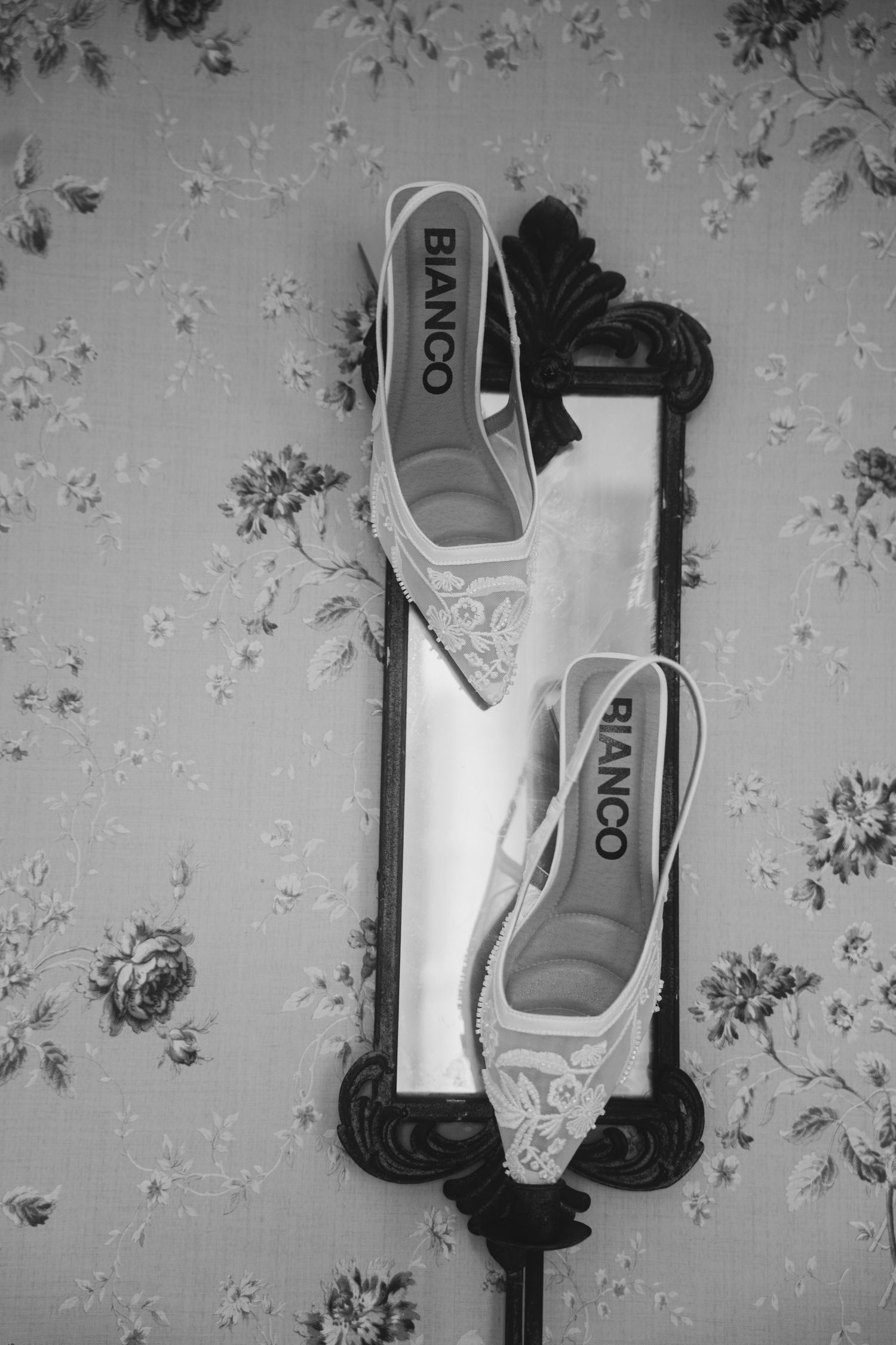 Black and white photo of two lace Bianco slingback bridal heels displayed on an ornate mirror against floral wallpaper.