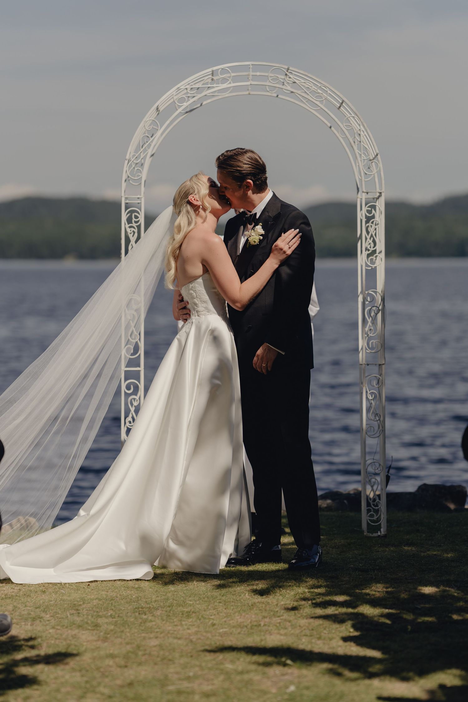 Bride in a white gown and long veil kisses her groom in a dark suit under a white arch beside a lake.