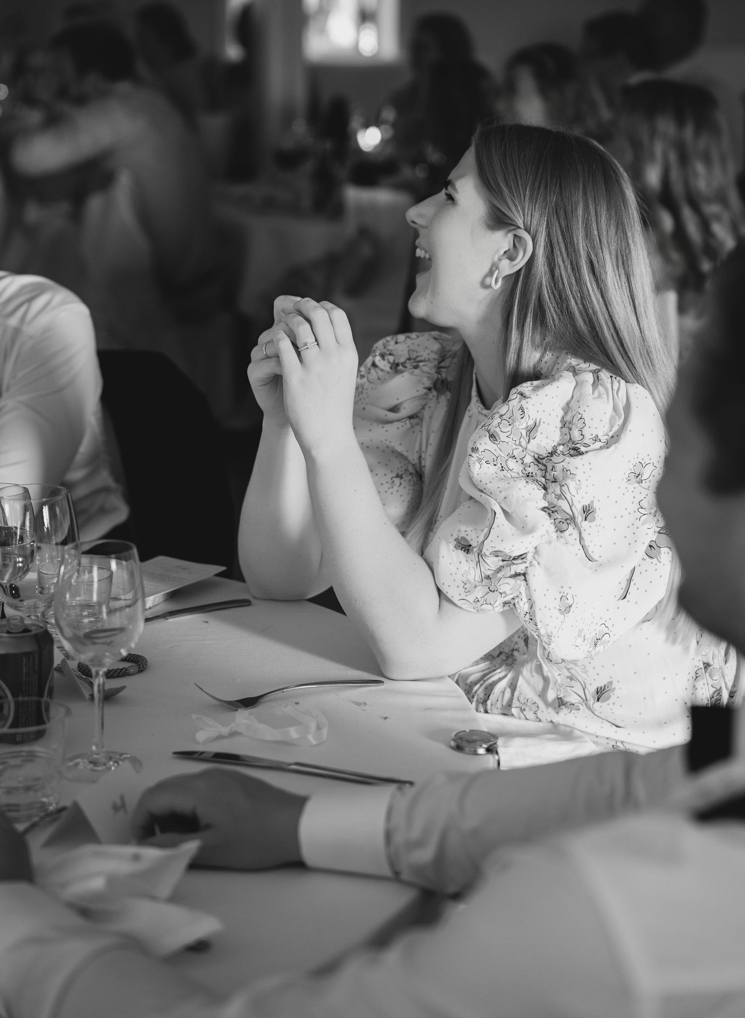 Black and white photo of a laughing woman in a floral dress clasping her hands at a wedding reception table.