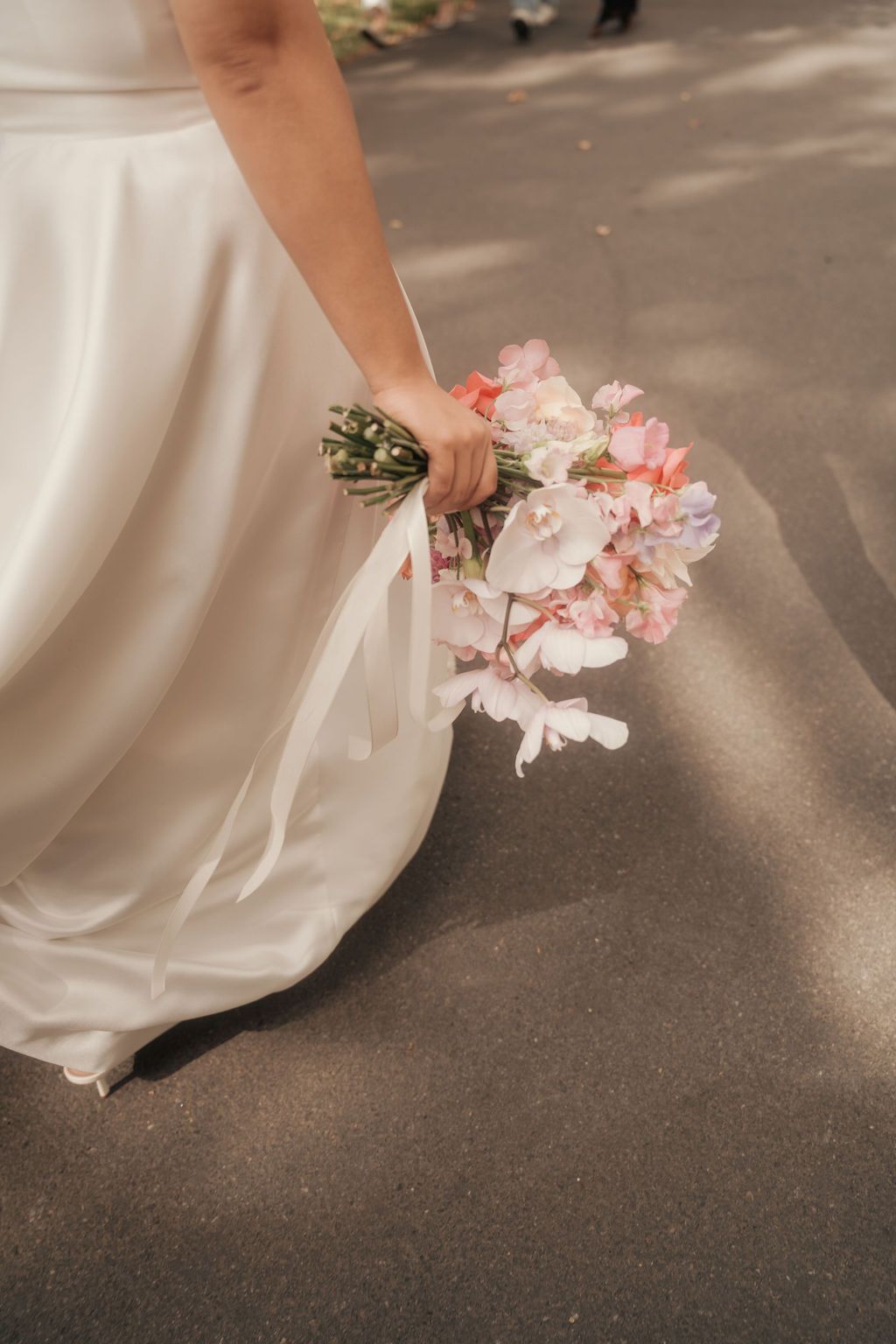 A person in a white wedding dress holds delicate pink flowers while standing on a dark floor.