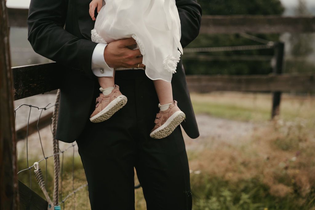 A person in a black suit holds a small child wearing white against a blurred outdoor background.