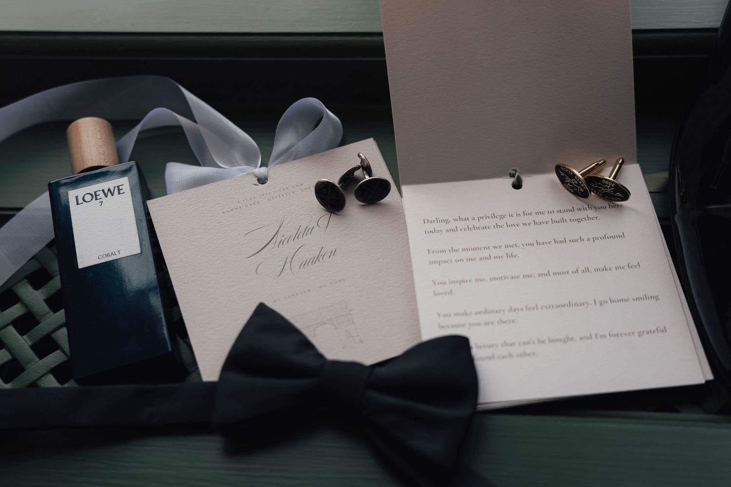 Elegant groom accessories flat lay featuring Loewe cologne, navy bow tie, cufflinks, and wedding invitation on a dark surface.