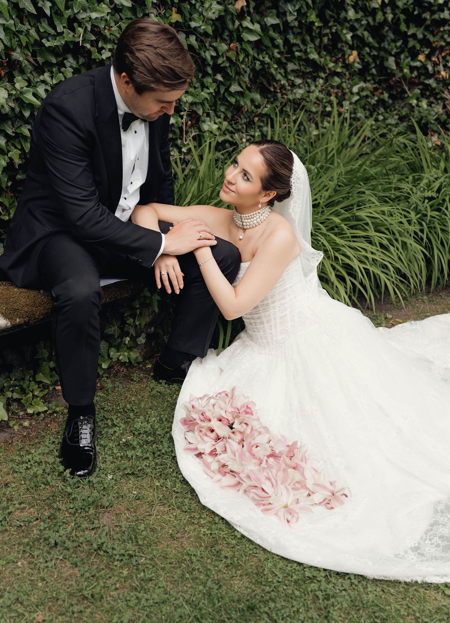 Bride in white gown with pink floral details sits on grass beside groom in black tuxedo at outdoor wedding.