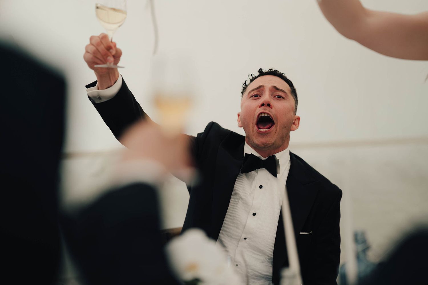 Man in tuxedo raising champagne glass, celebrating joyfully at a formal wedding reception event.