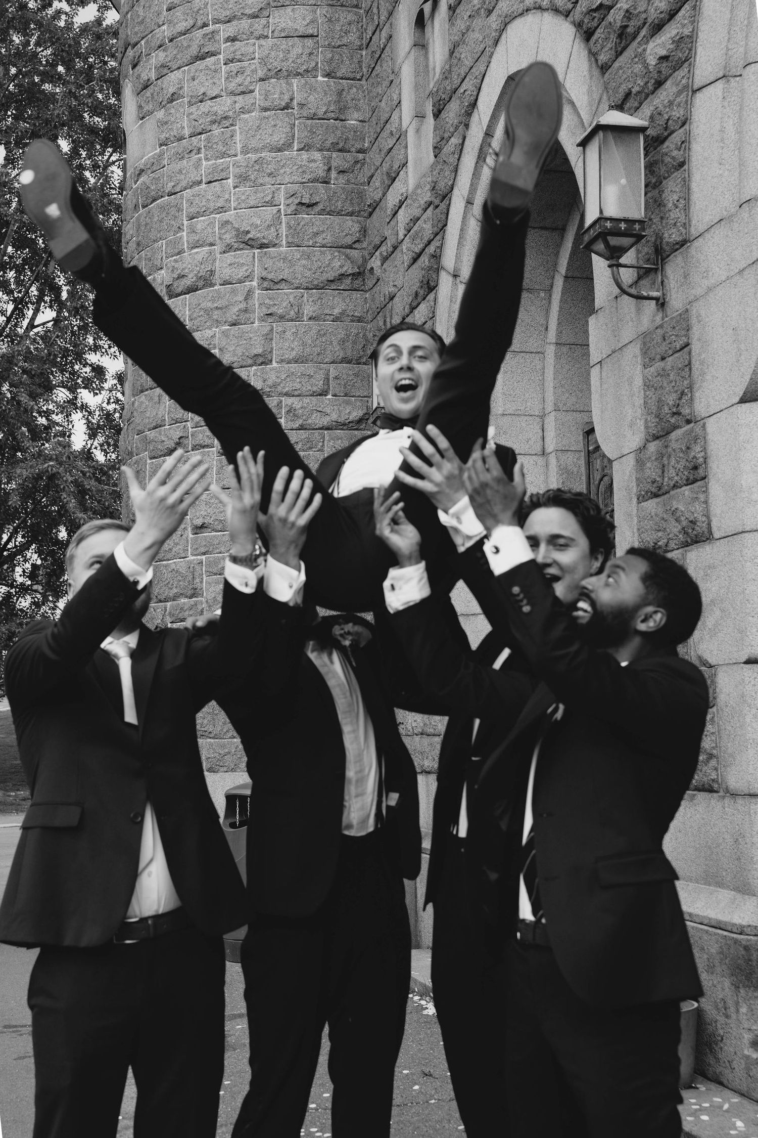 Groomsmen lifting groom in the air celebrating outside stone church in black and white wedding photo.