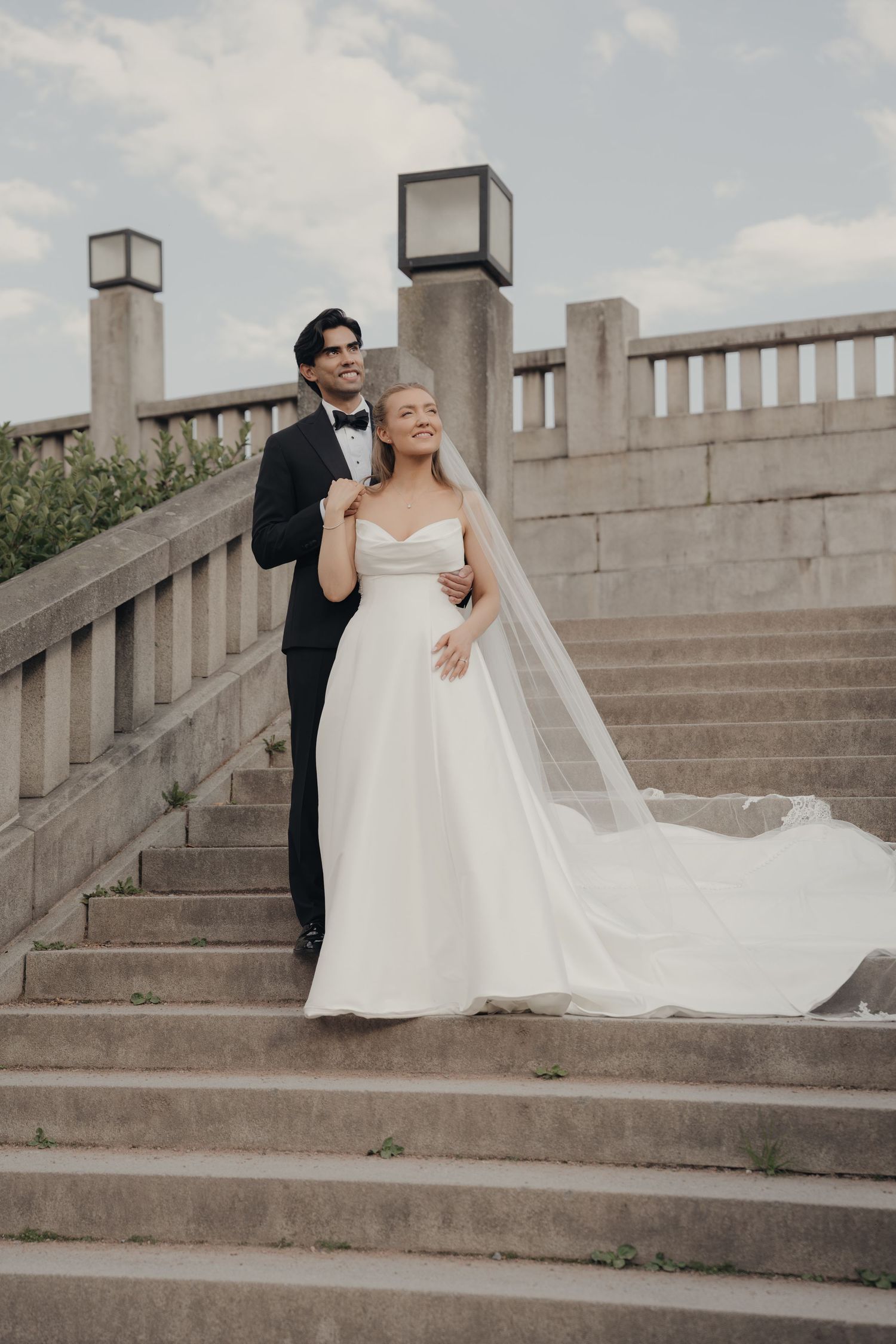 Bride in a strapless white gown and long veil stands embraced by her groom in a tuxedo on stone steps for an outdoor wedding portrait.